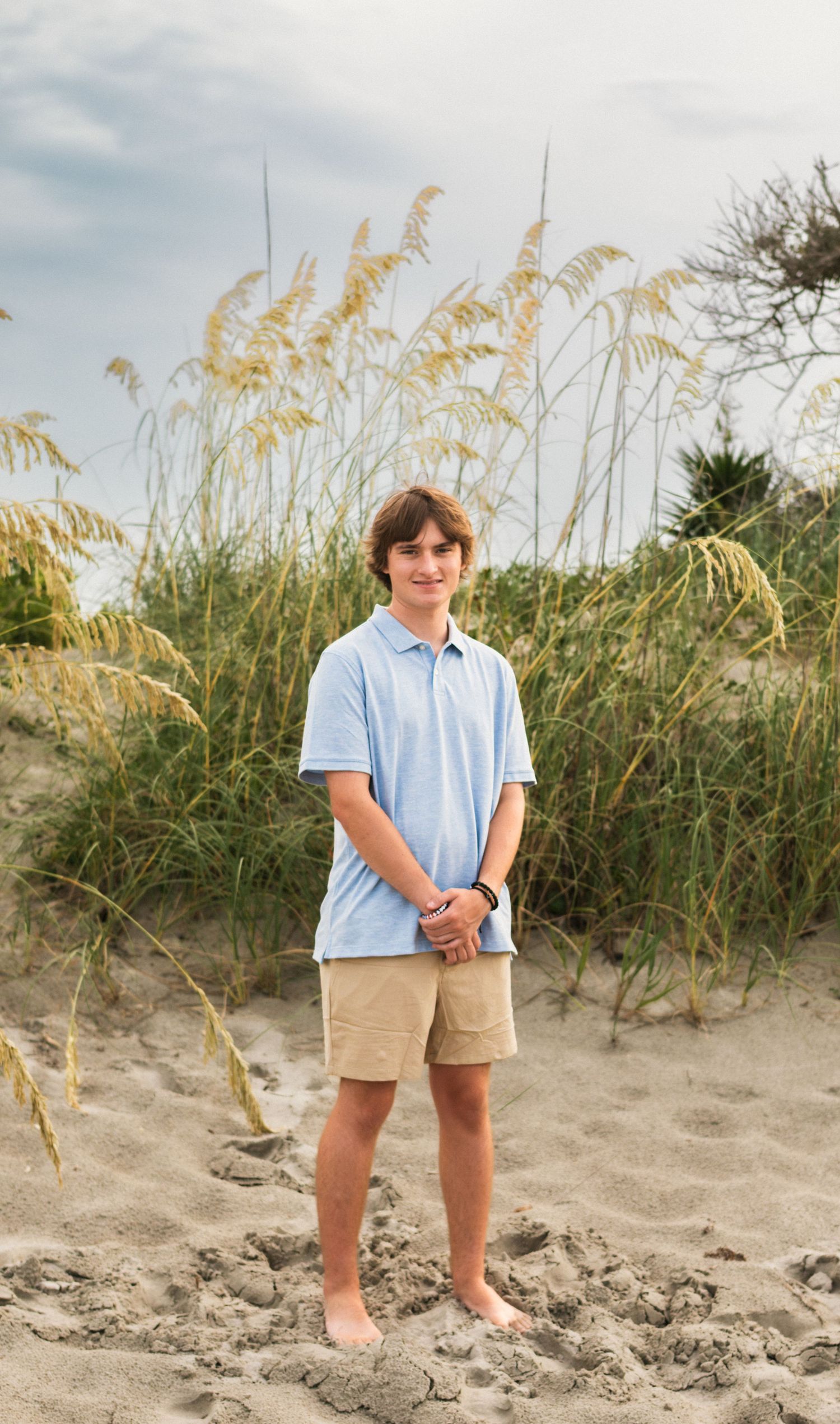 A teenage boy in a light blue polo shirt and khaki shorts stands barefoot on sandy beach dunes with sea oats behind him.