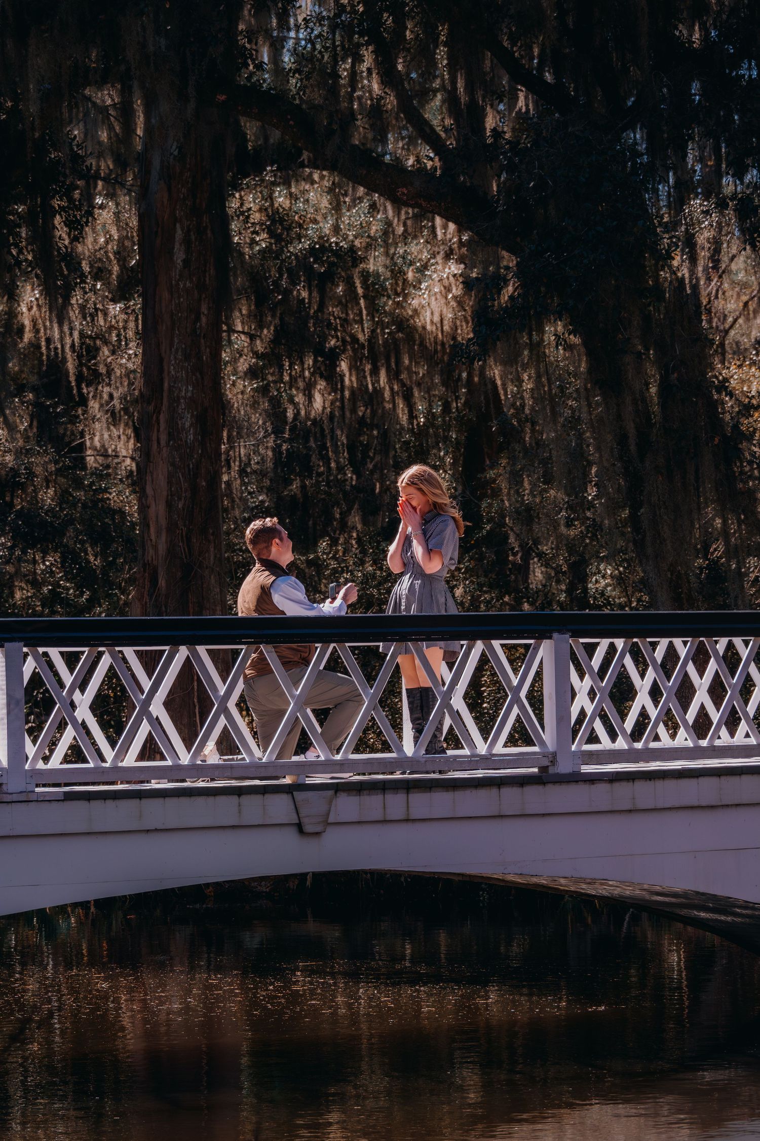 Man kneeling in proposal on white bridge over pond surrounded by moss-draped trees as woman covers her mouth in surprise.