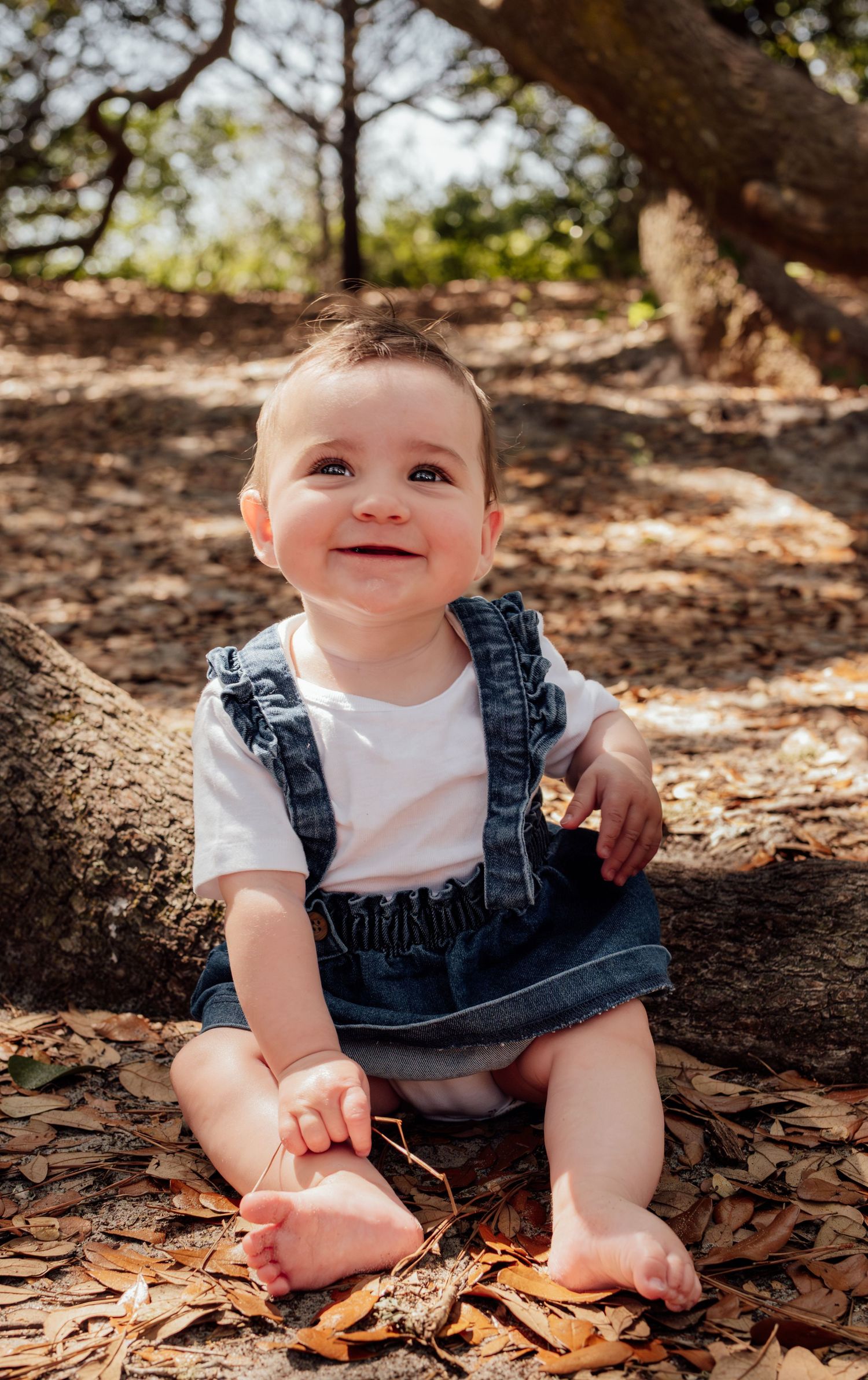 Smiling baby in denim overalls sitting barefoot on fallen leaves under a tree outdoors.