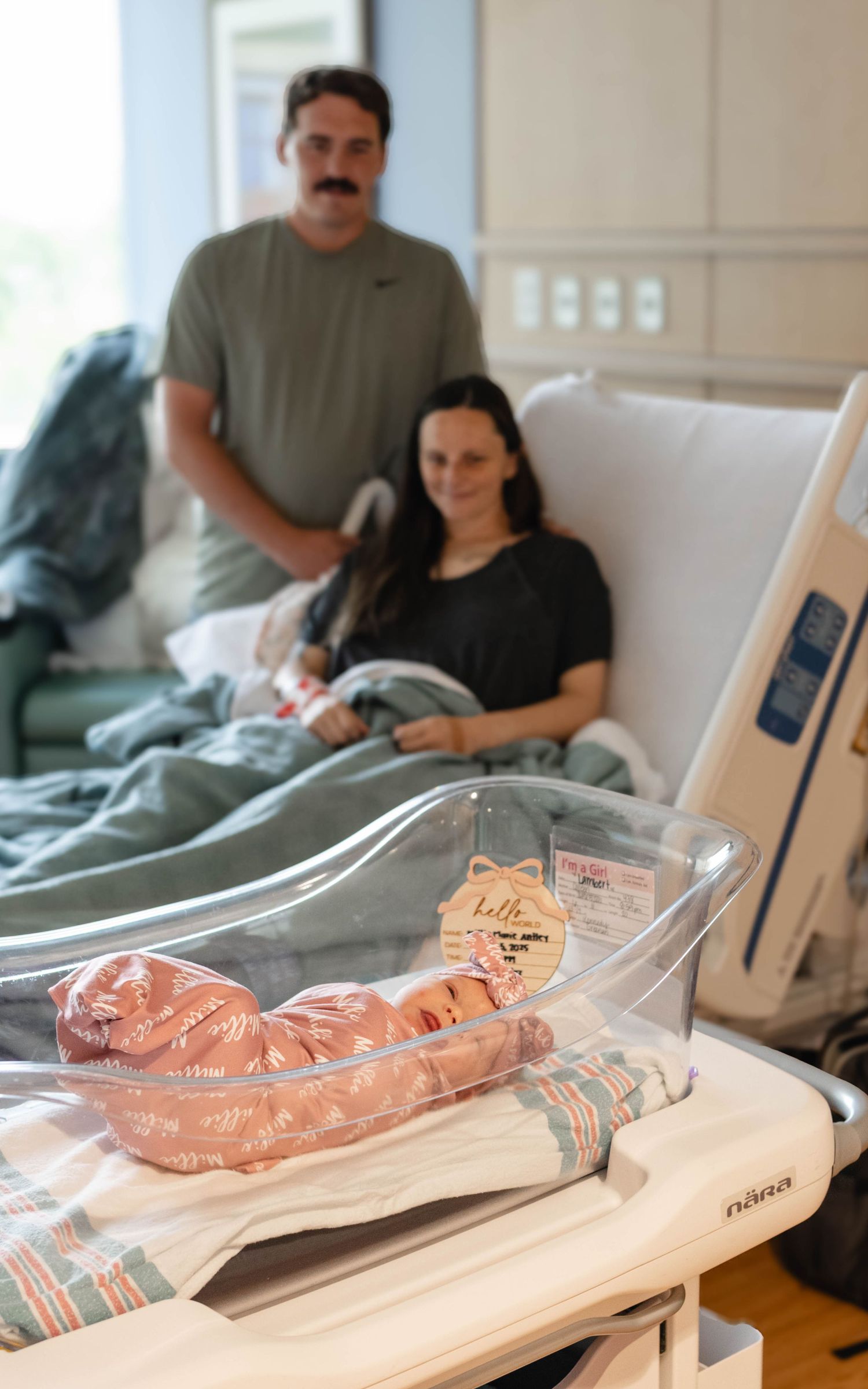 Newborn baby in hospital bassinet with proud parents resting in background after delivery.