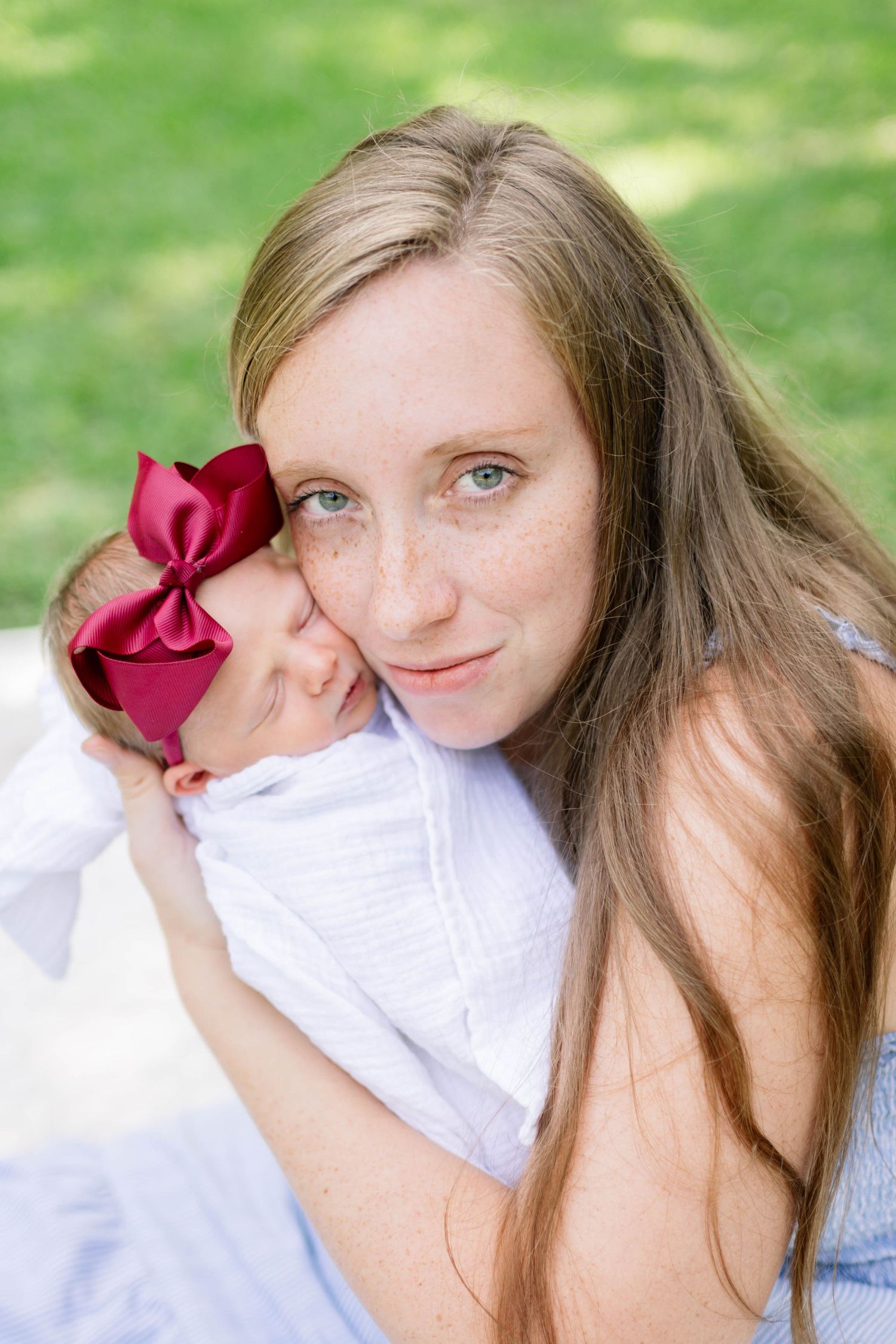 A young mother with long hair and freckles tenderly holds her sleeping baby girl wearing a red bow headband outdoors.