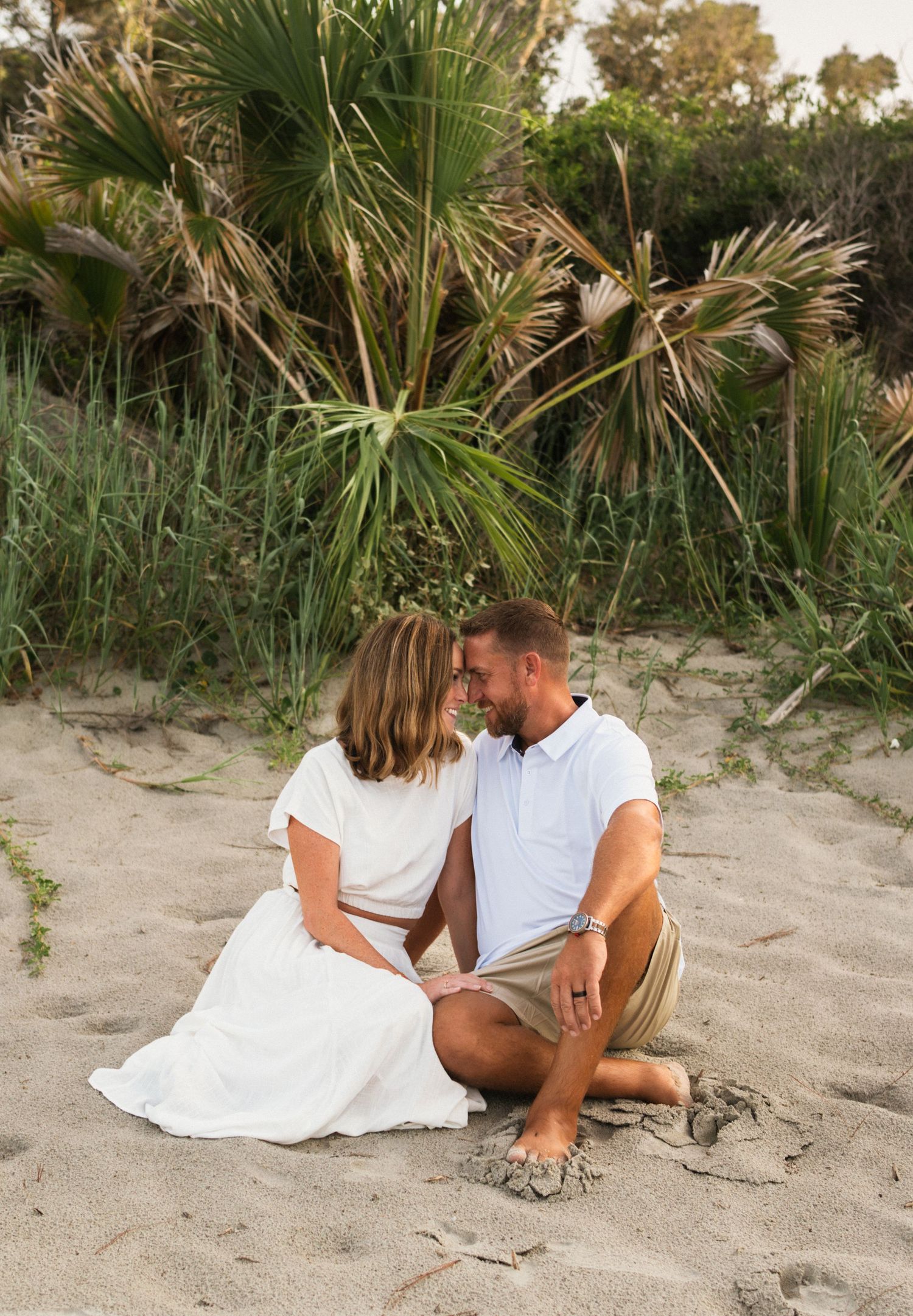 Couple sitting on sandy beach in white outfits, sharing a romantic moment with tropical palms in background.