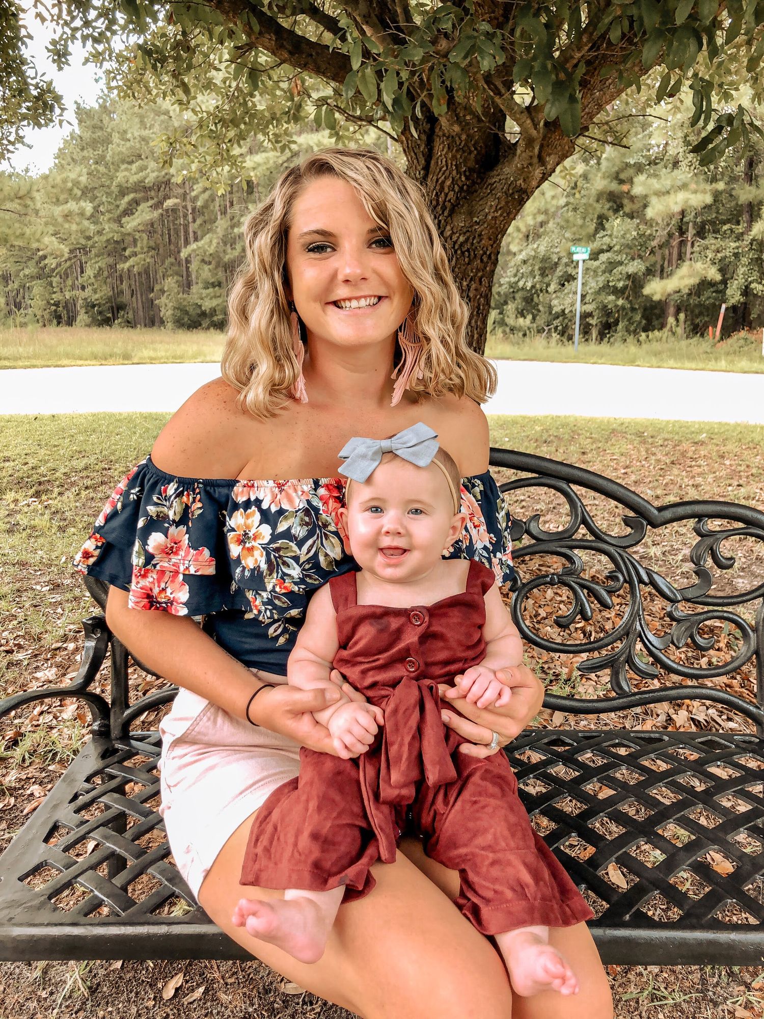 A smiling young mother sits on an ornate bench holding her baby girl in a red romper outdoors.