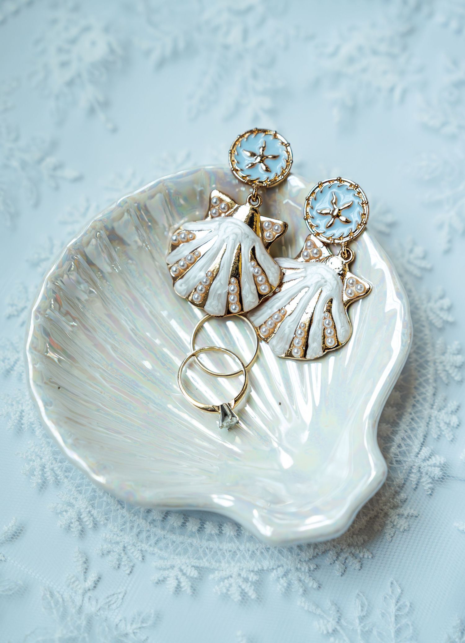 White shell-shaped dish with gold and pearl seashell earrings and delicate rings on a light blue background.