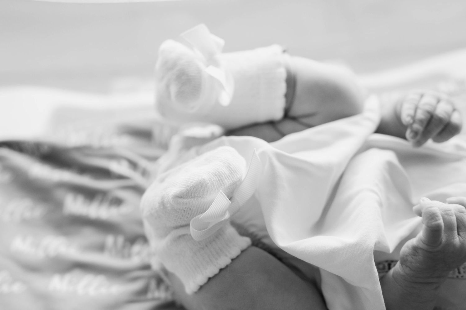 Black and white photo of a newborn baby's tiny feet and hands wearing white socks, lying on a soft blanket.