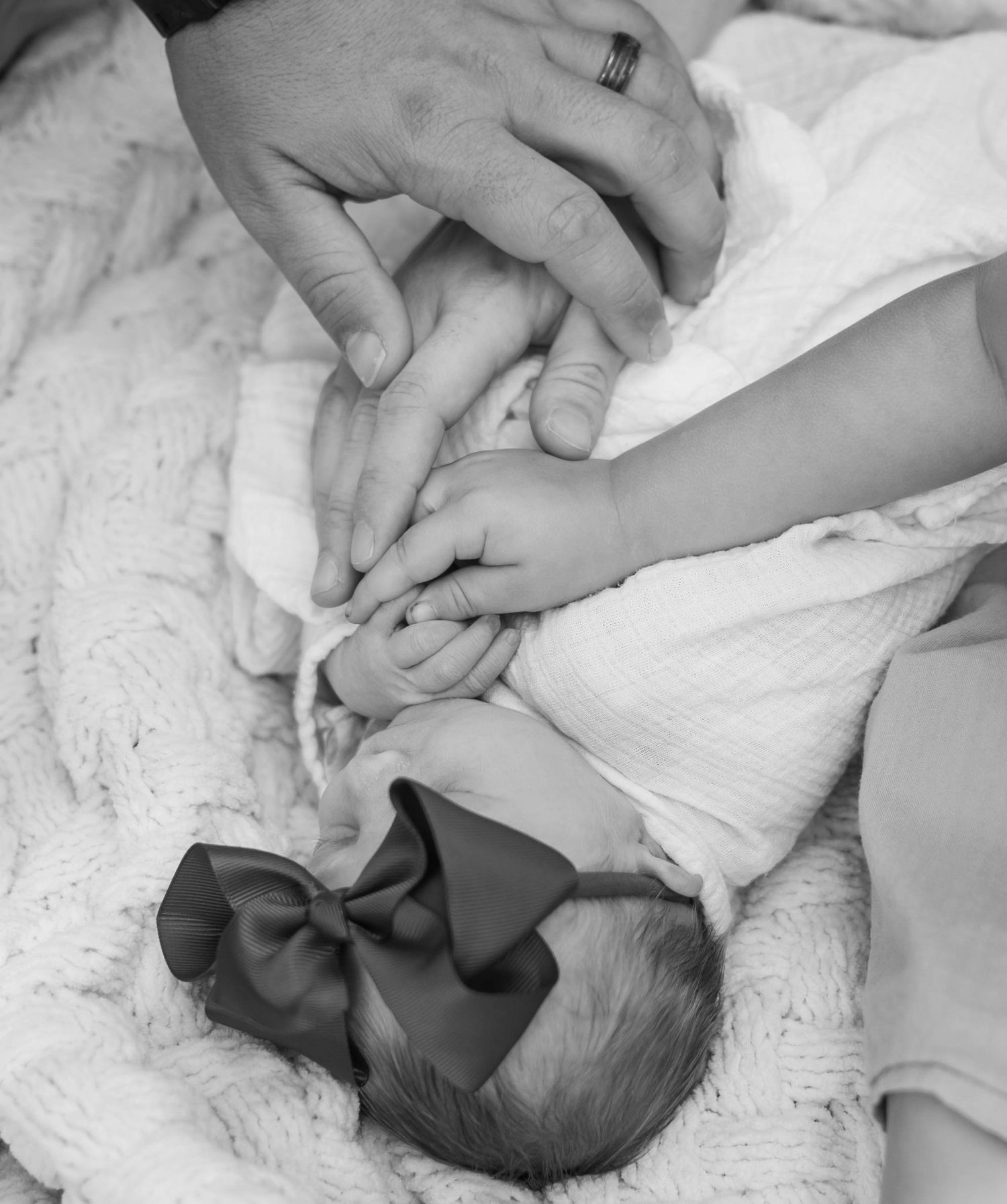 Black and white photo of newborn baby girl with bow being held by parents hands.