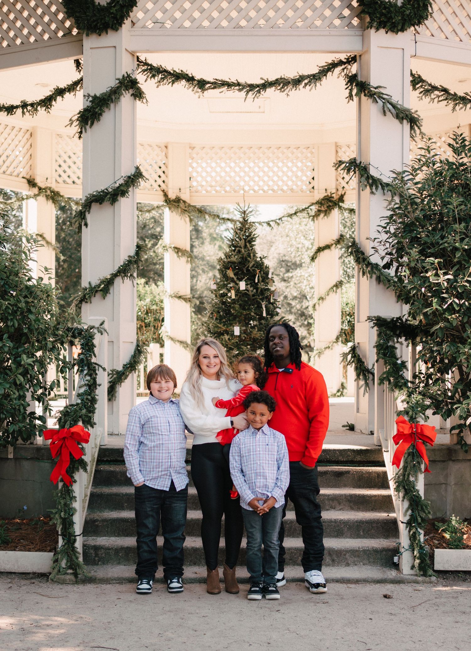 Family of five posing outdoors at Christmas-decorated gazebo with garland, red bows, and holiday tree backdrop.