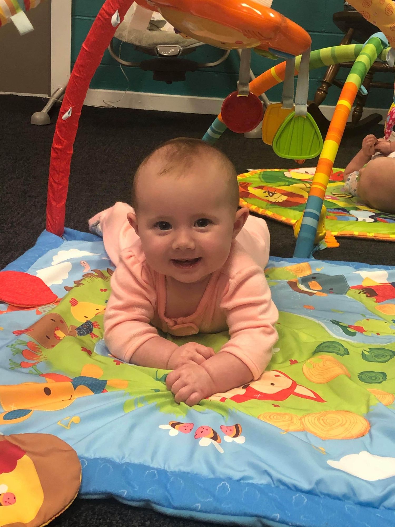 A happy baby girl in a pink outfit enjoys tummy time on a colorful Winnie the Pooh play mat.