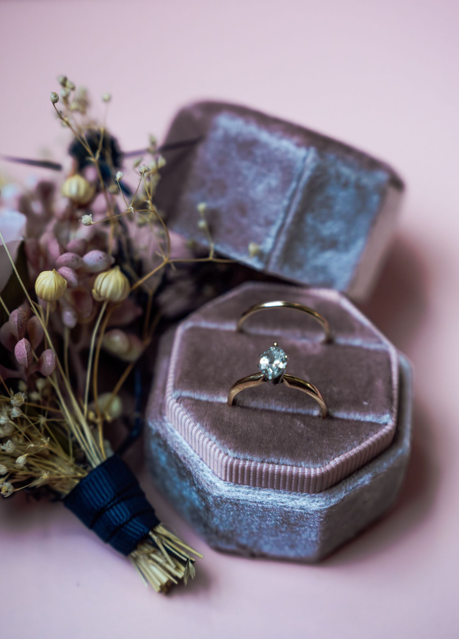 Gold solitaire engagement ring displayed in a velvet hexagonal ring box beside dried flowers on a pink background.
