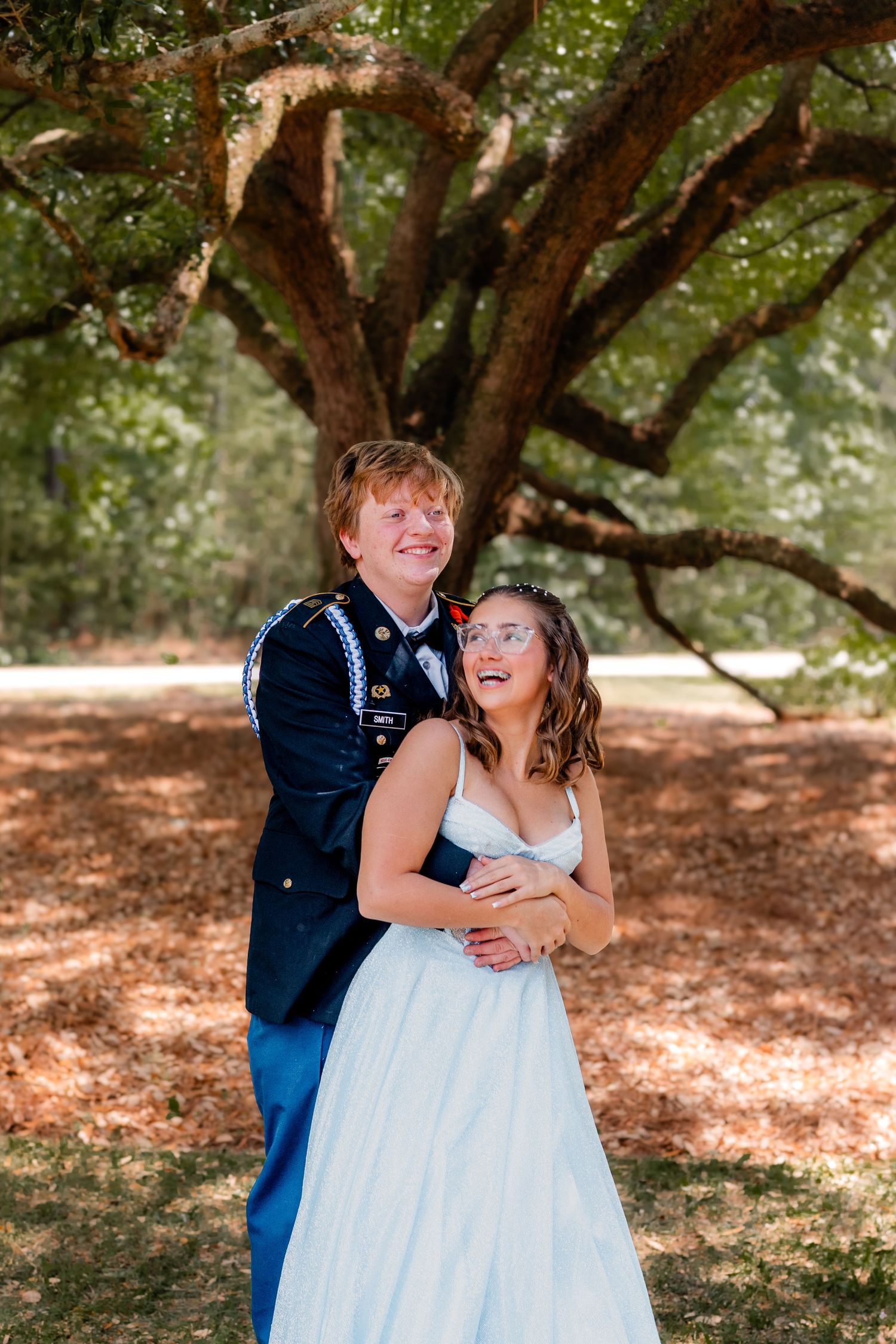 Smiling couple embracing outdoors, man in military dress blues hugging woman in blue gown.