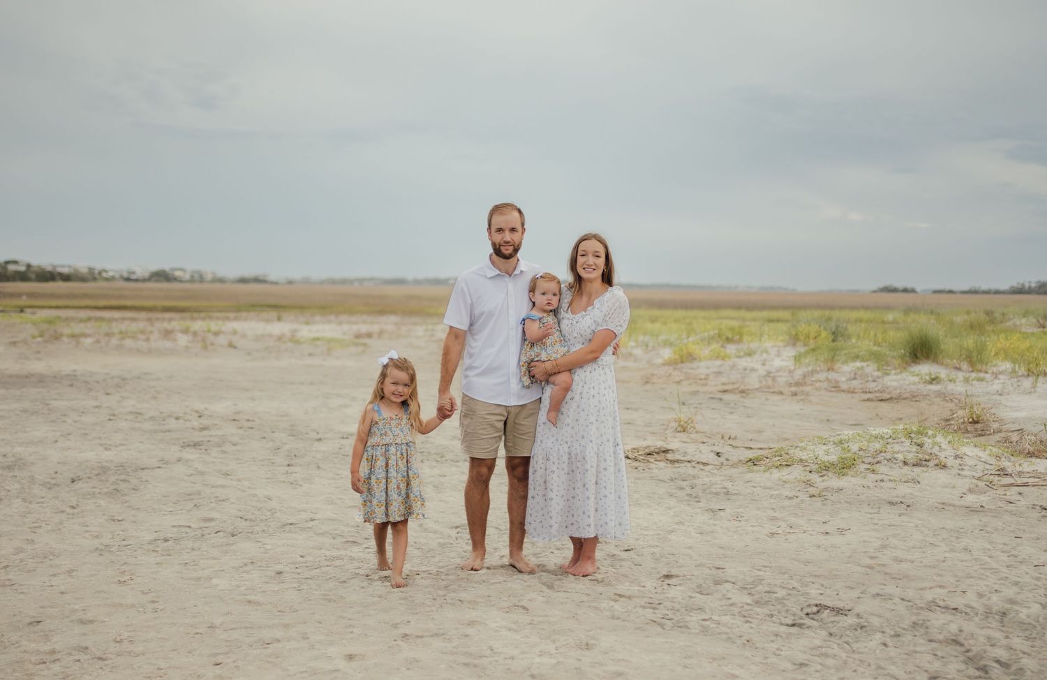 Family of four posing on sandy beach, parents with two young children, overcast sky and marsh in background.