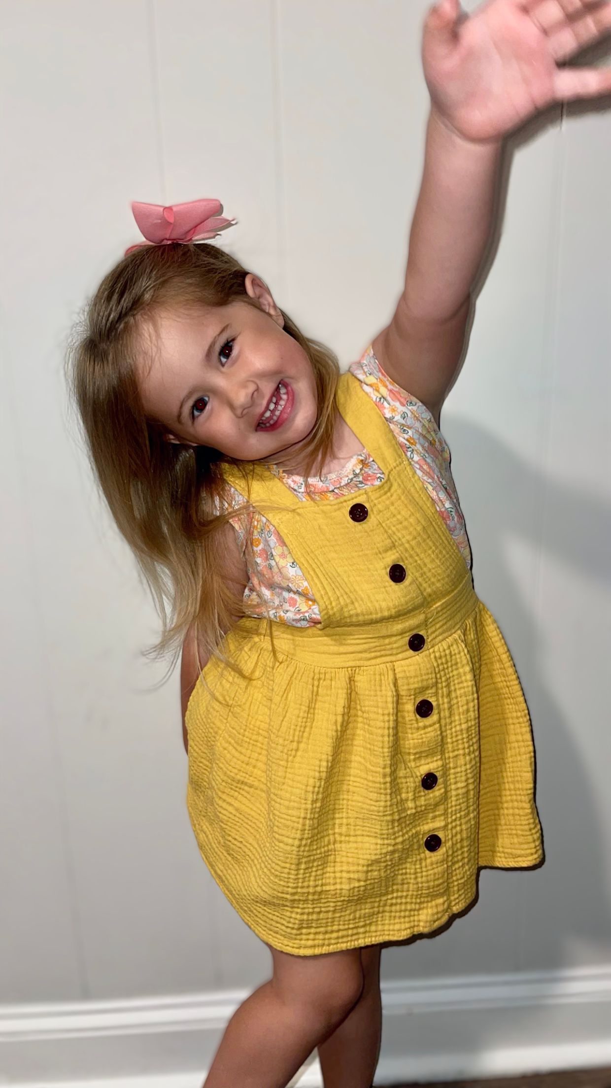 A cheerful little girl in a yellow jumper and floral top raises her arm and smiles against a white wall.