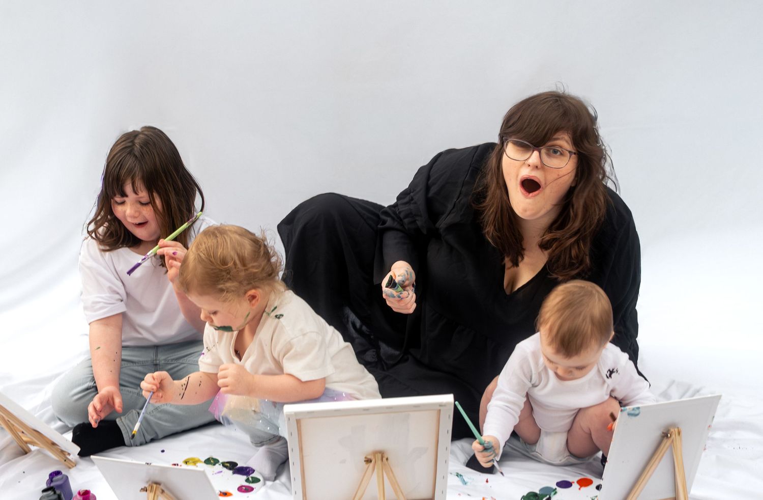 Woman with three young children painting on canvas during a creative art session on white backdrop.