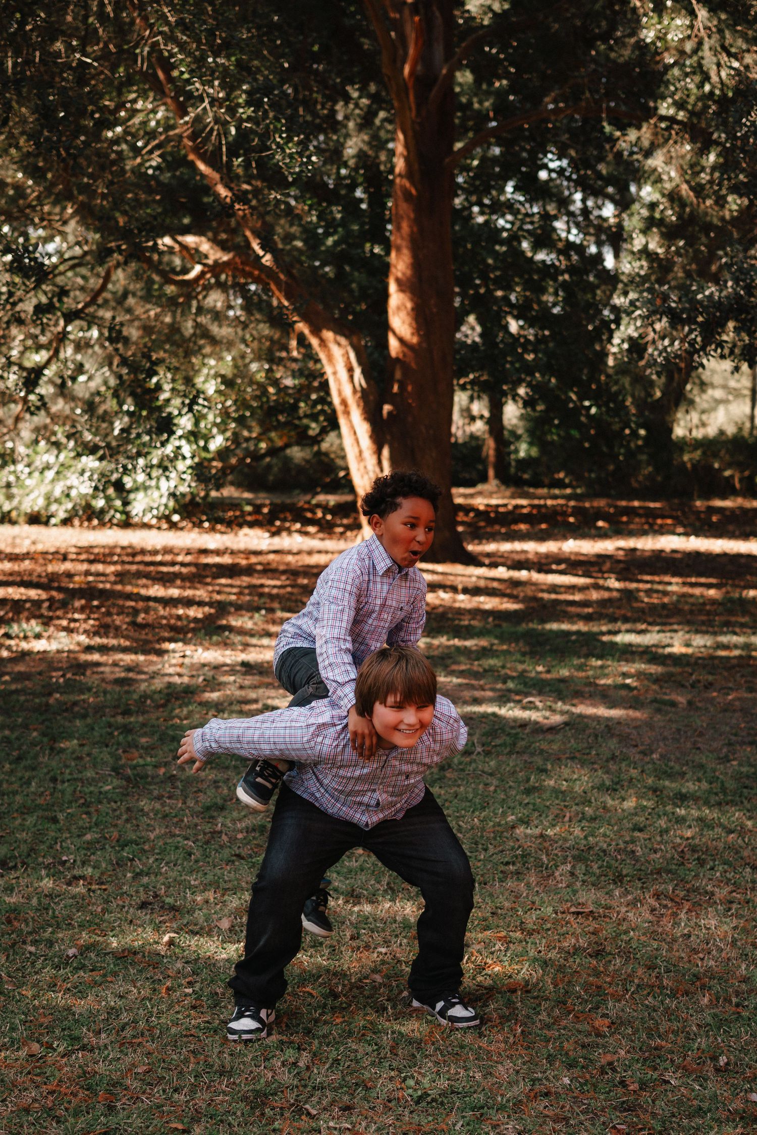 Two boys playing piggyback ride in a sunny park with large trees and green grass in the background.