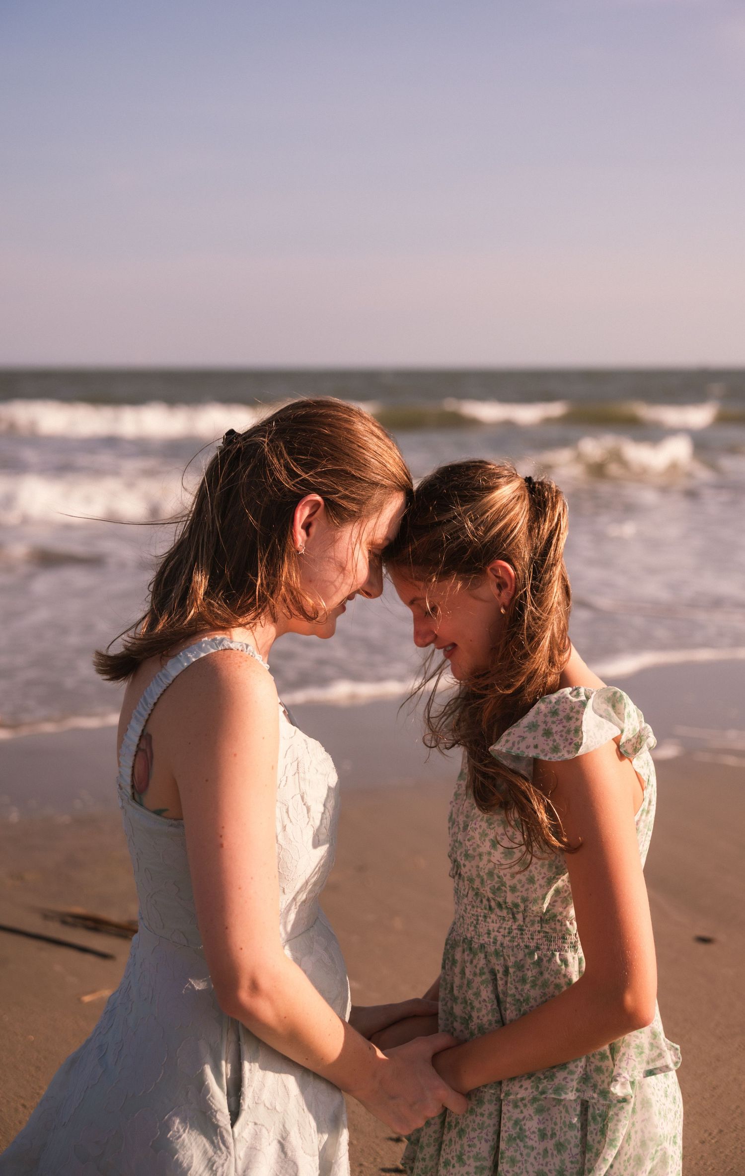Mother and daughter in floral dresses hold hands on a sandy beach, touching foreheads with ocean waves behind them.