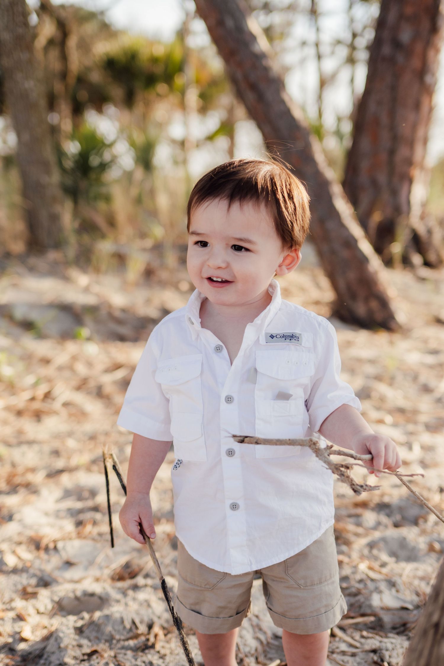 Toddler boy in white Columbia shirt and khaki shorts holding a stick outdoors among trees.