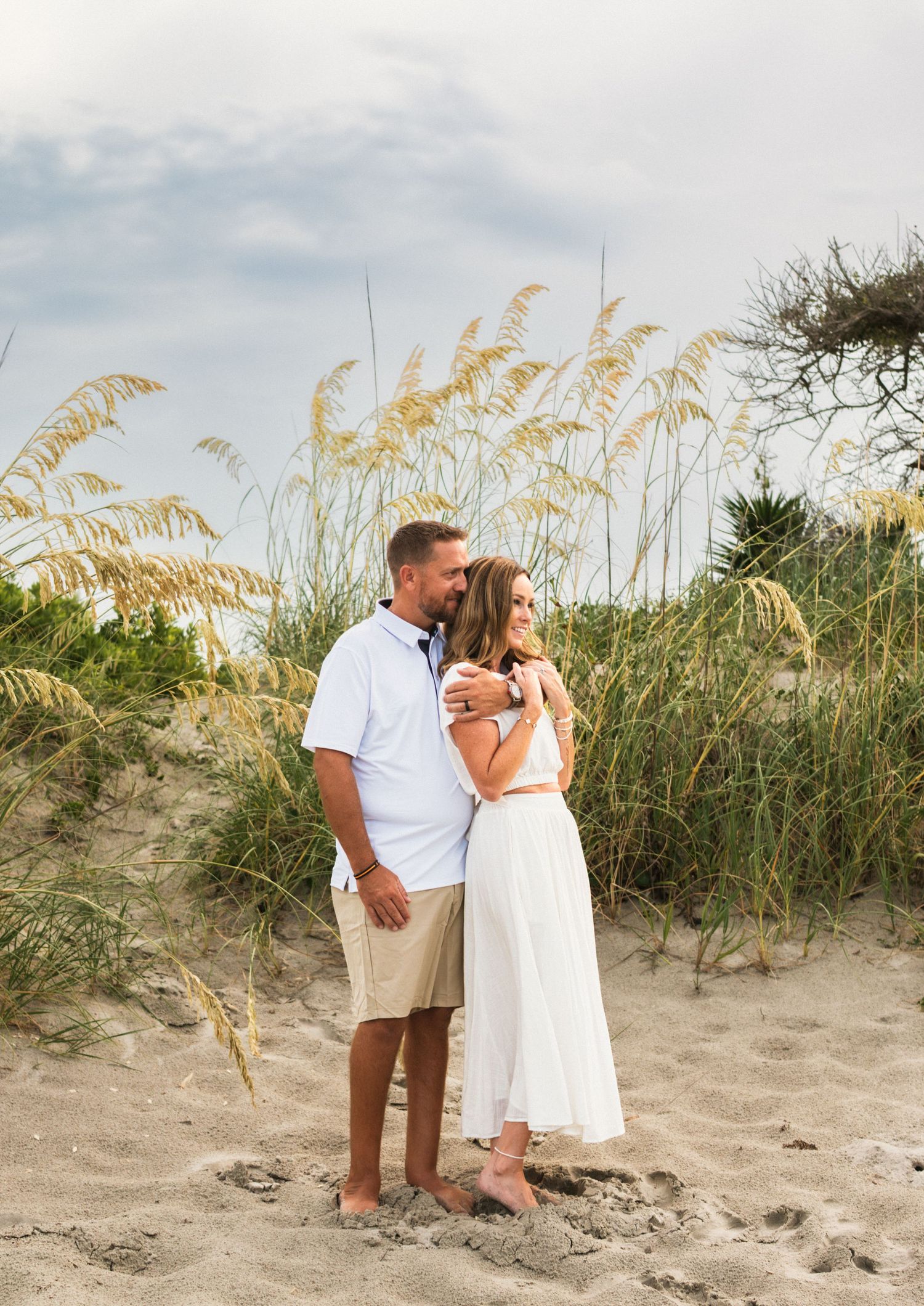 Couple embracing barefoot on sandy beach dunes with sea oats, man in white shirt and khakis, woman in white dress.