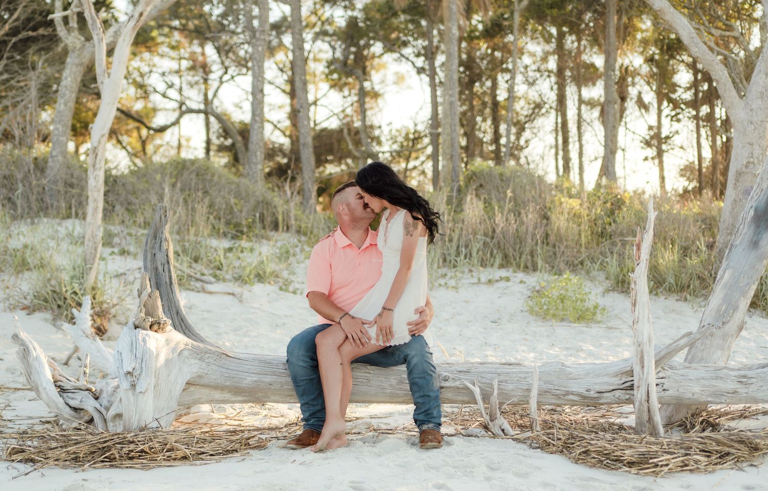 Couple sharing a romantic moment on a sandy beach, sitting on driftwood surrounded by trees at golden hour.