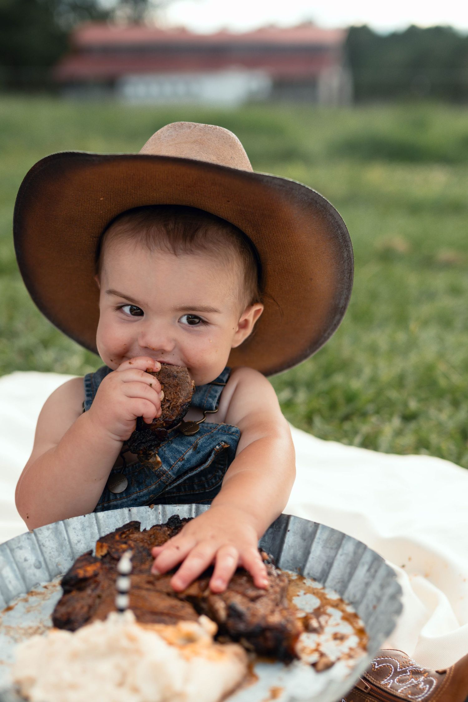 Baby in cowboy hat eating cake smash outdoors on white blanket in green field, wearing denim overalls.