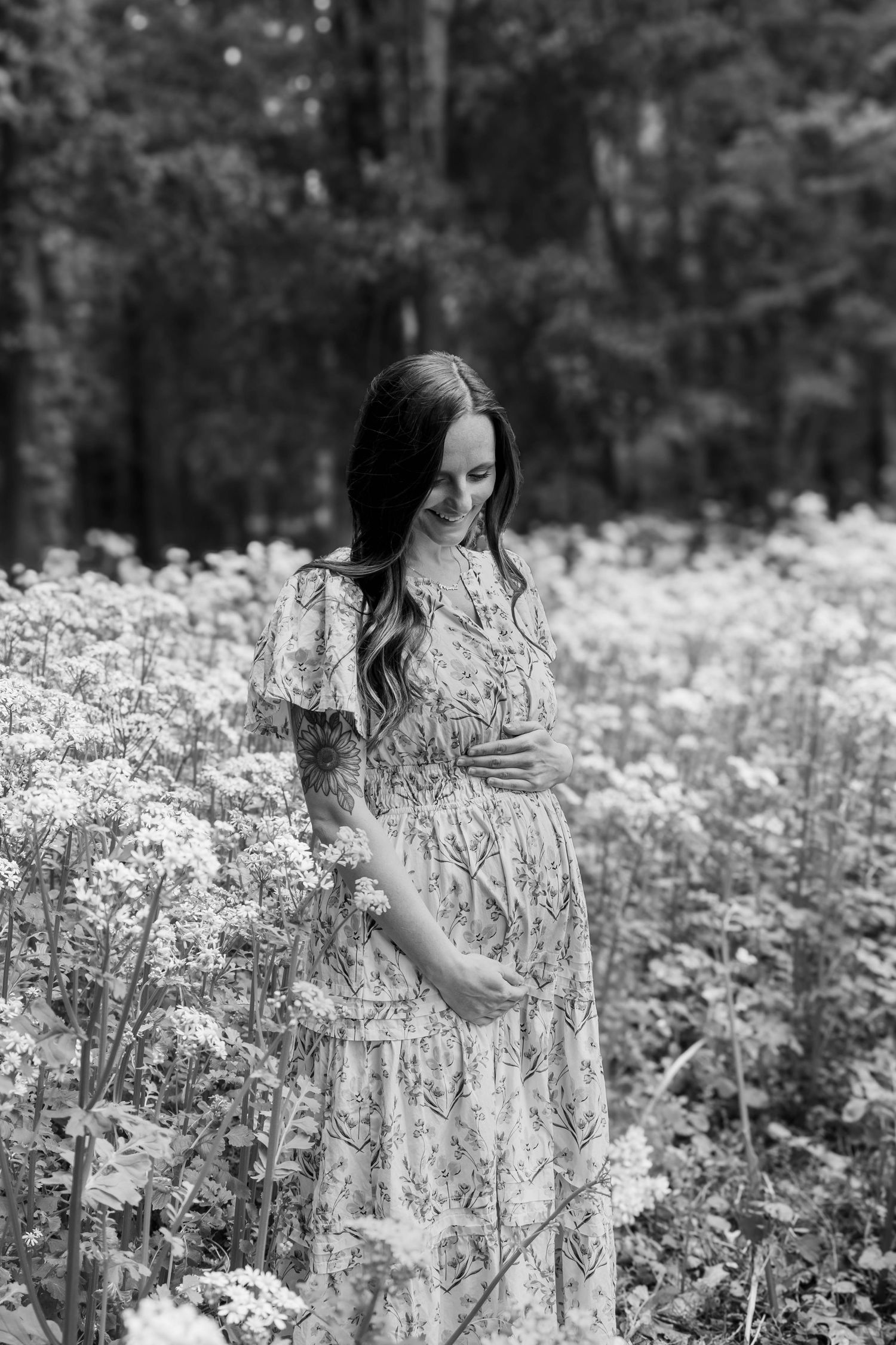 Black and white photo of a smiling pregnant woman in a floral dress standing among wildflowers in a forest.