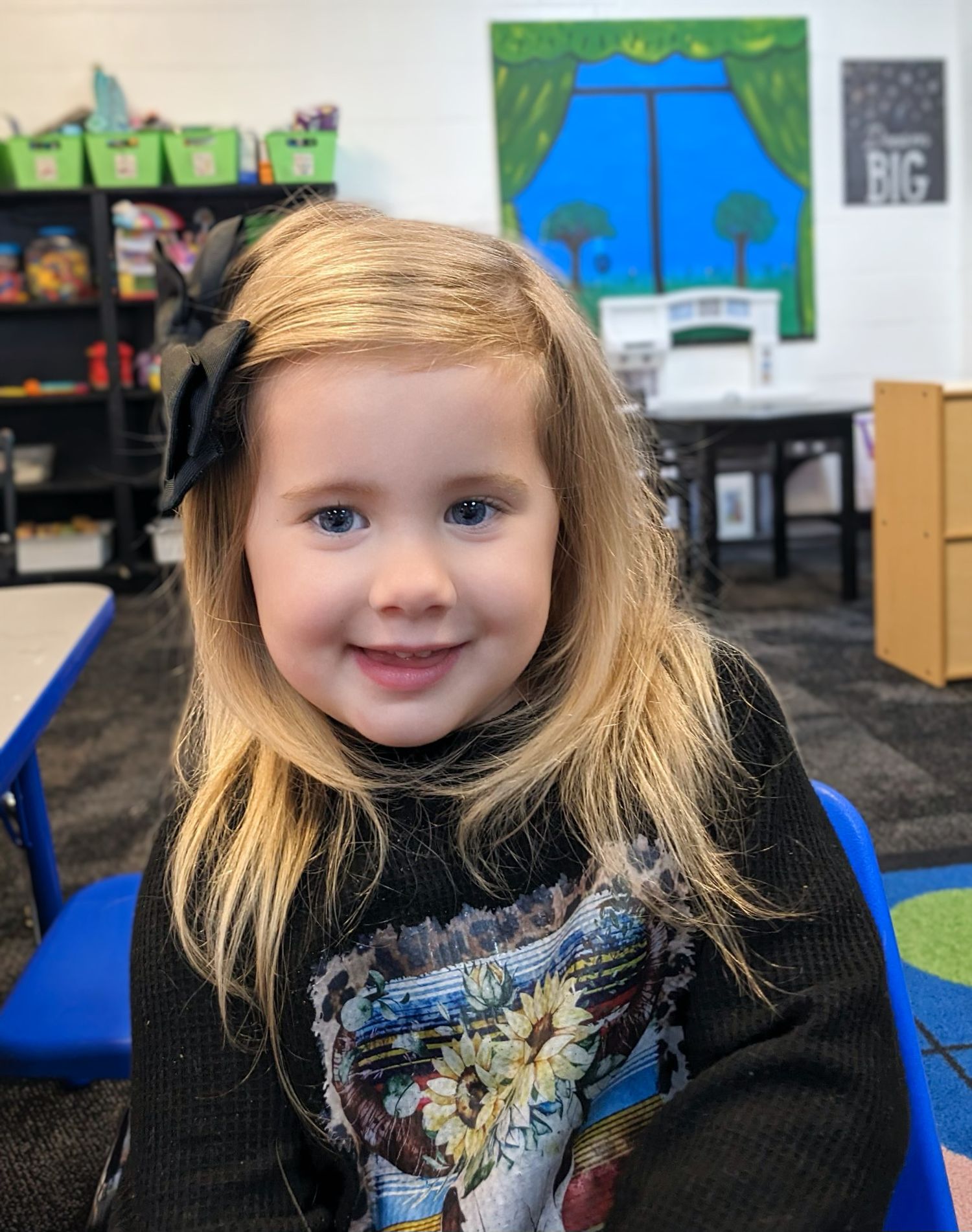 Smiling blonde girl with black bow in hair wearing graphic tee in colorful classroom setting.