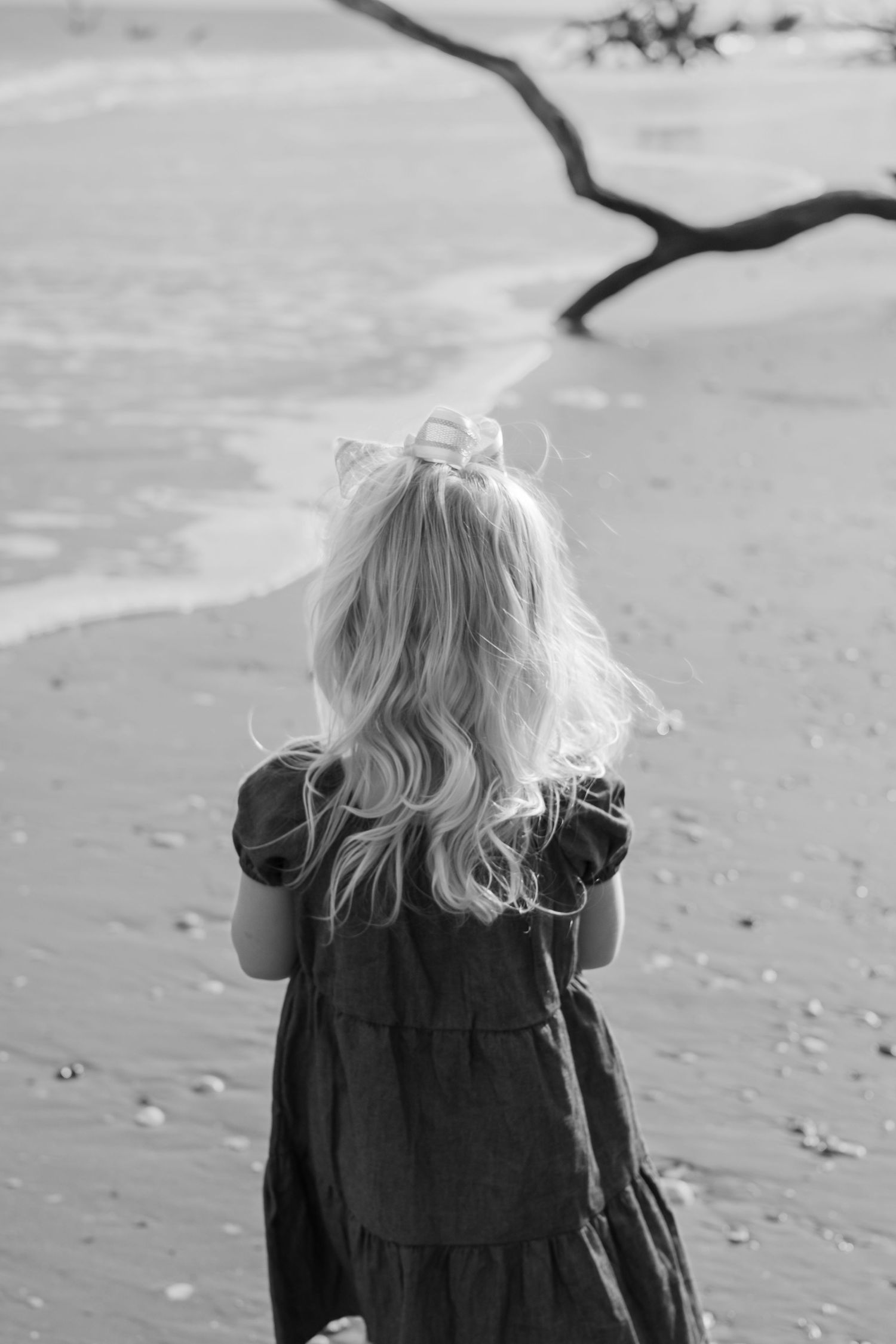 Black and white photo of a young blonde girl in a dress standing on a beach facing the ocean waves.