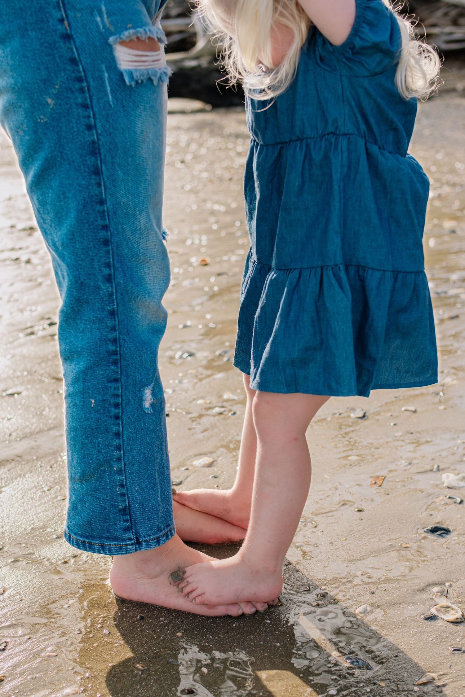 Barefoot child in blue dress standing on sandy beach beside adult in ripped jeans, enjoying a seaside moment together.