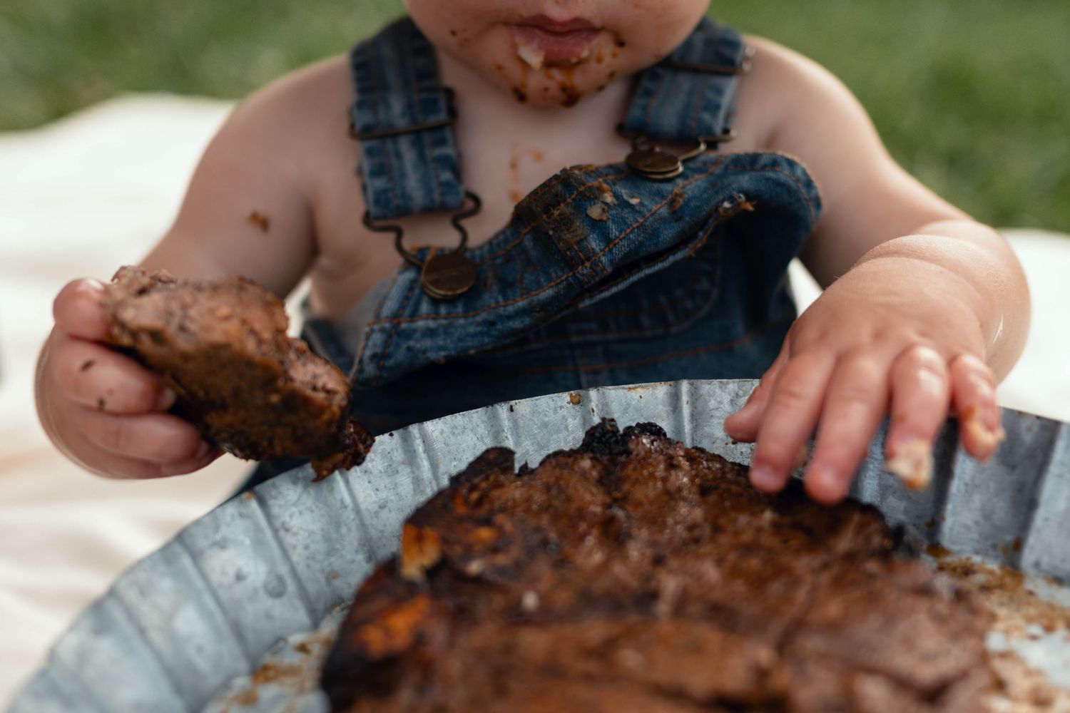 Messy toddler in denim overalls eating chocolate cake with hands during first birthday smash cake celebration outdoors.