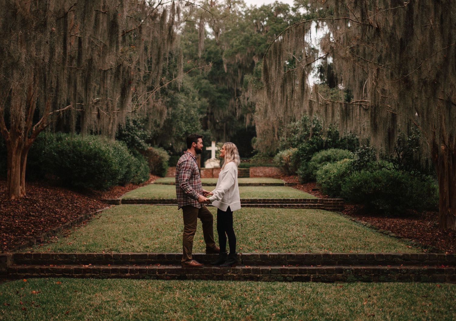 Couple holding hands in a moss-draped garden pathway surrounded by ancient oak trees.