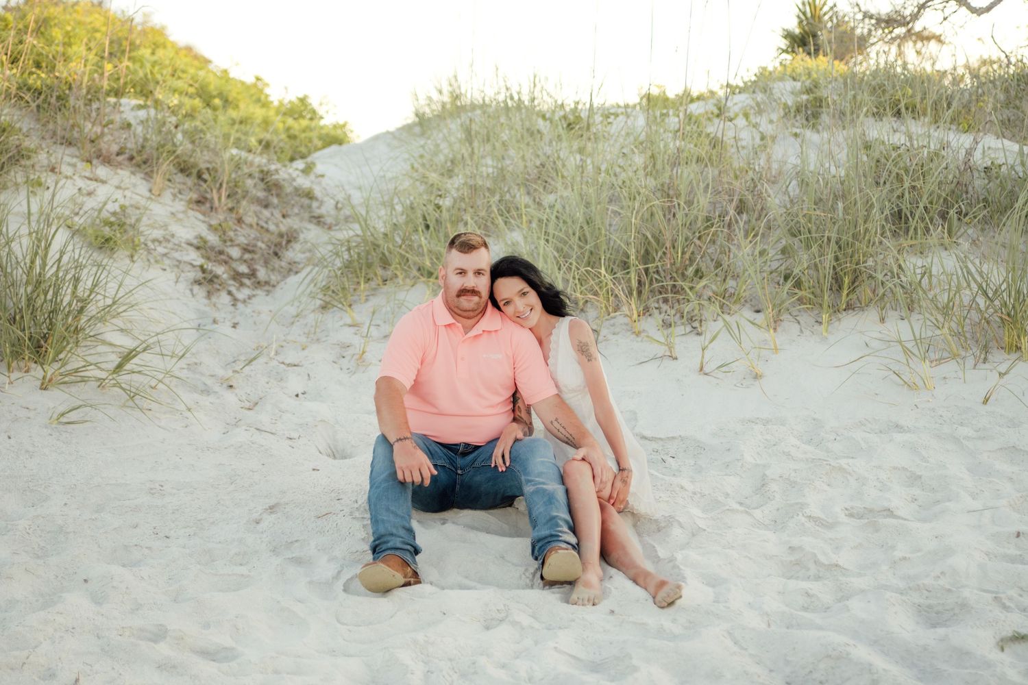 Couple sitting together on sandy beach dunes with sea grass, man in pink shirt and jeans, woman in beige dress.