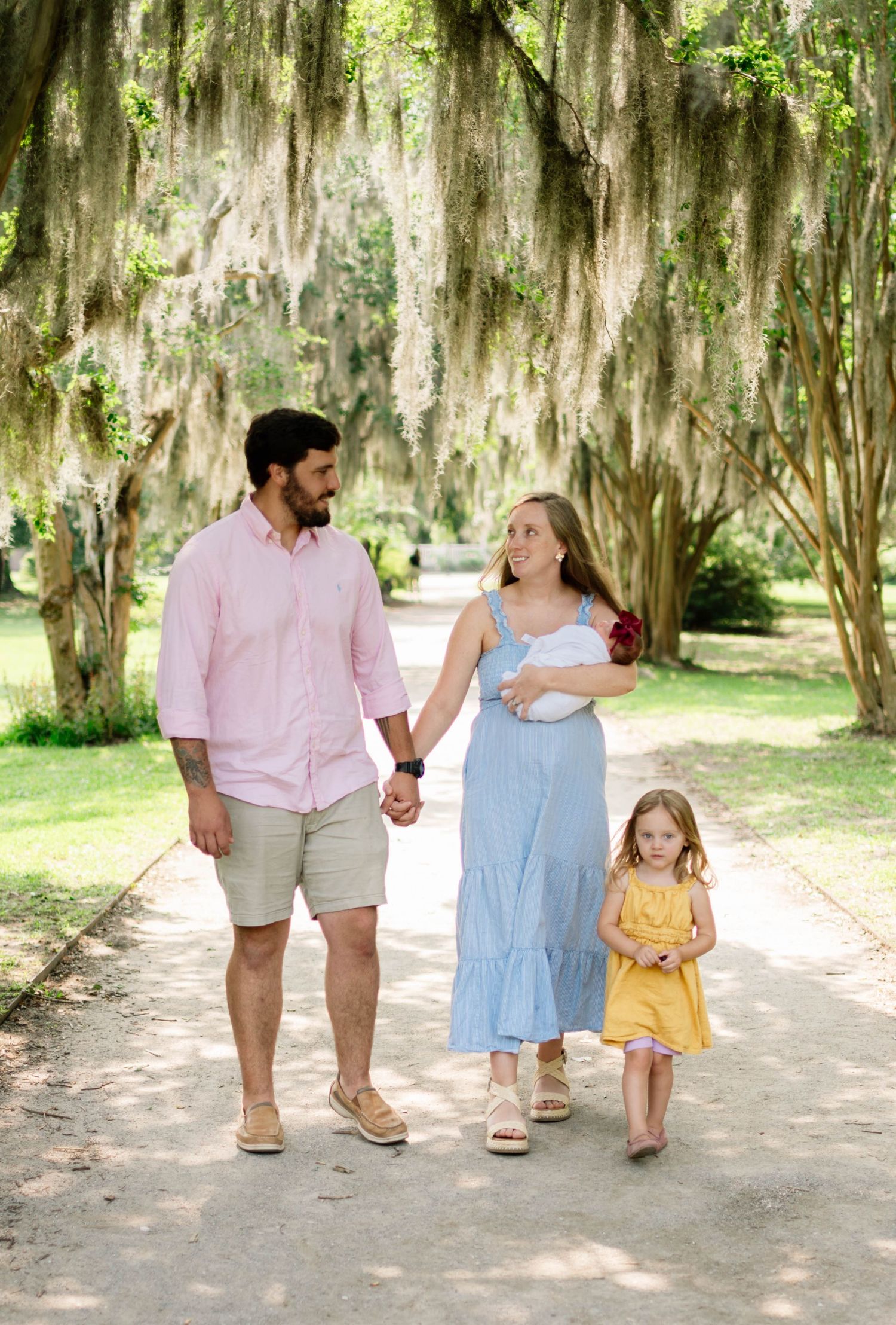 Family of three walking hand-in-hand on a shaded path under Spanish moss-draped oak trees.