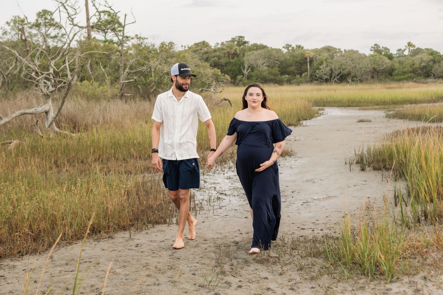 Pregnant couple holds hands walking on sandy marsh path surrounded by tall grass during outdoor maternity photo session.