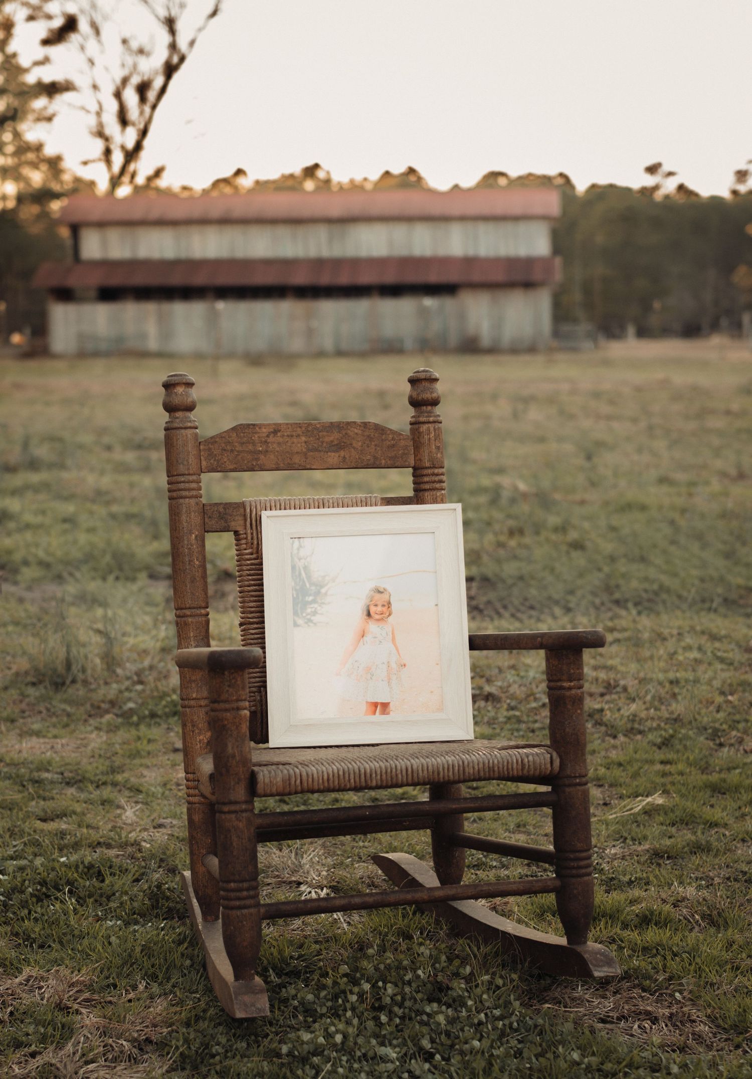 Vintage wooden rocking chair holding a framed child portrait in an open field near old barn.