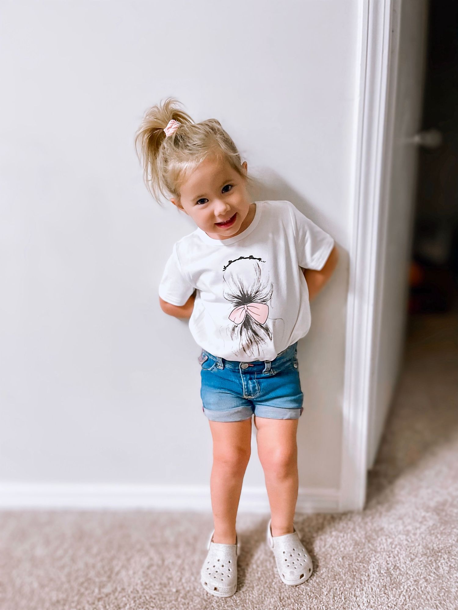 Cute blonde toddler girl in white floral tee and denim shorts posing by white wall.