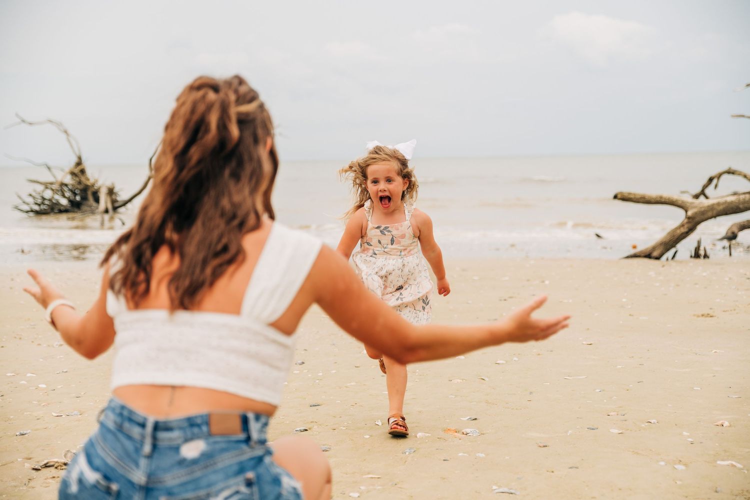 A laughing little girl runs with arms wide open toward a woman on a sandy beach with driftwood in the background.
