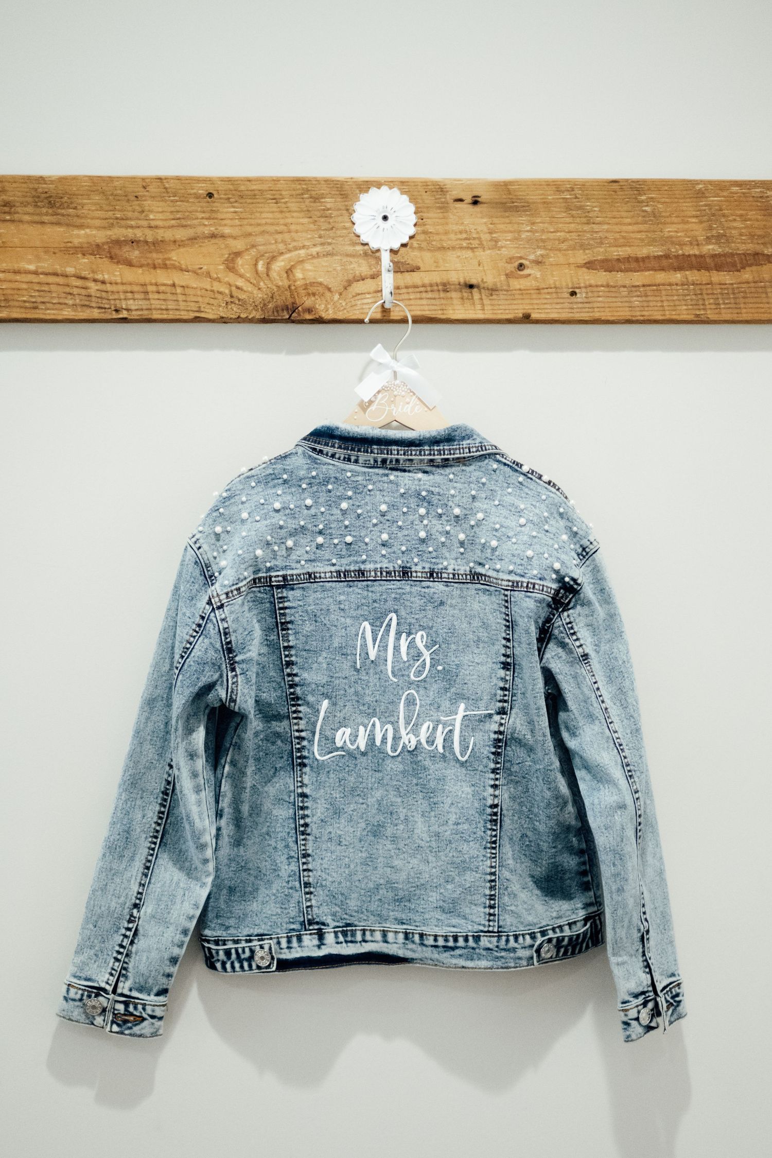 Personalized denim jacket with pearl embellishments hanging on wooden rack, reading Mrs. Lambert, perfect bridal gift.