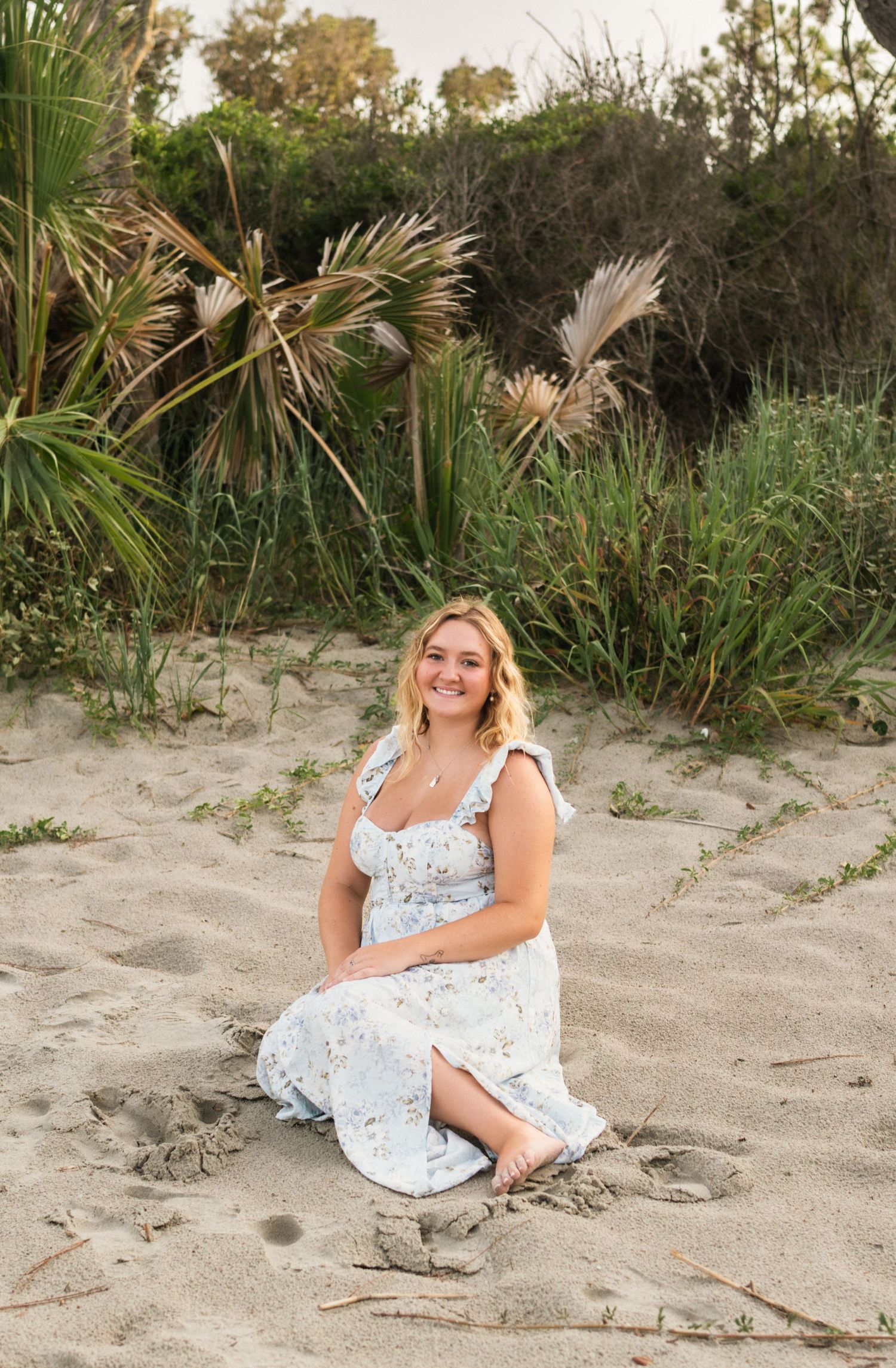 Young blonde woman in floral sundress sitting on sandy beach with palm trees and green foliage in background.