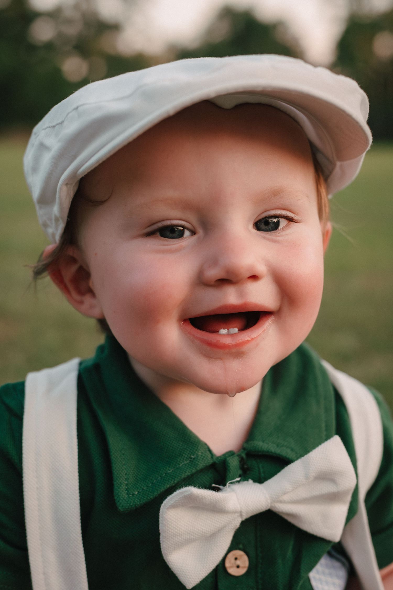 Smiling toddler boy wearing a white flat cap, green shirt, bow tie, and suspenders outdoors.