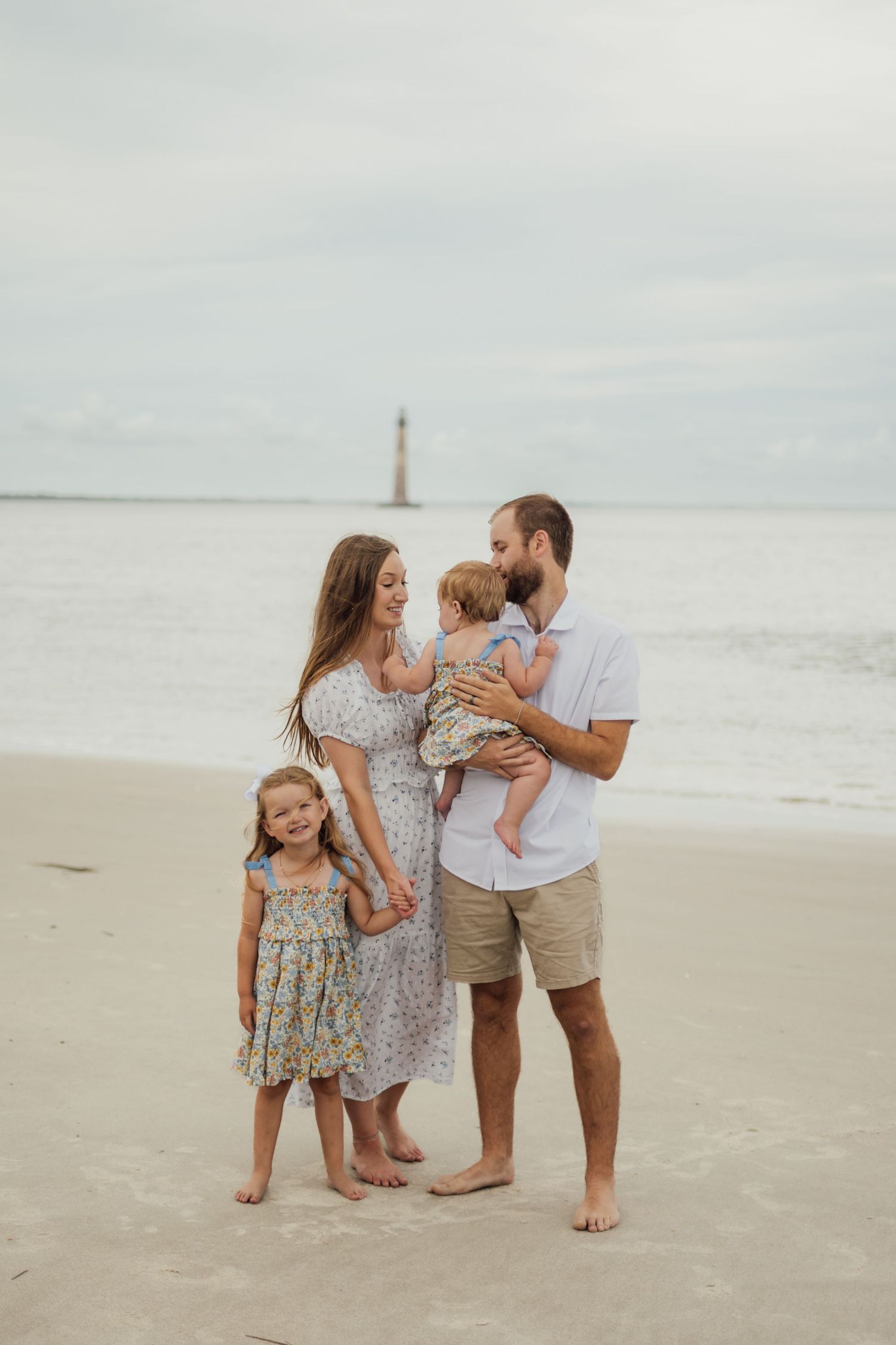 Family of four posing on sandy beach with lighthouse in background, parents holding children in matching floral dresses.