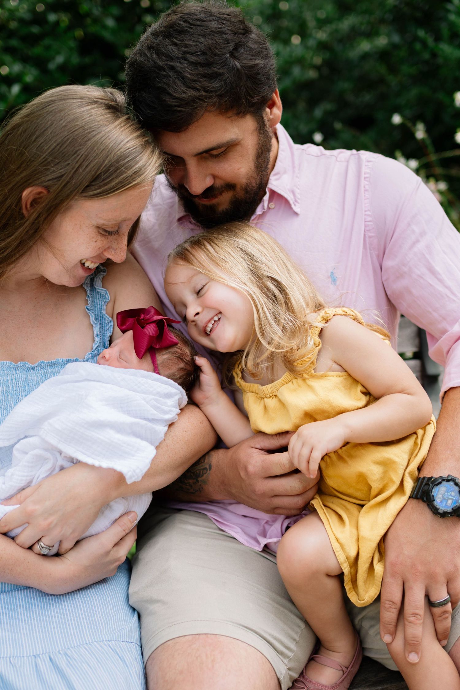 A happy family of four cuddles outdoors, parents smiling at newborn baby while toddler girl in yellow dress laughs joyfully.