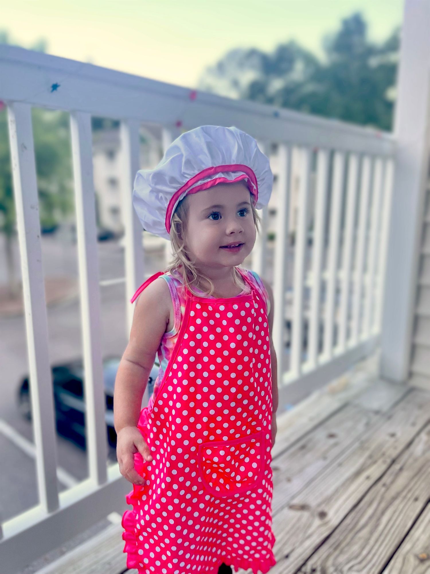 Smiling toddler girl in pink polka dot dress and white chef hat on wooden deck.