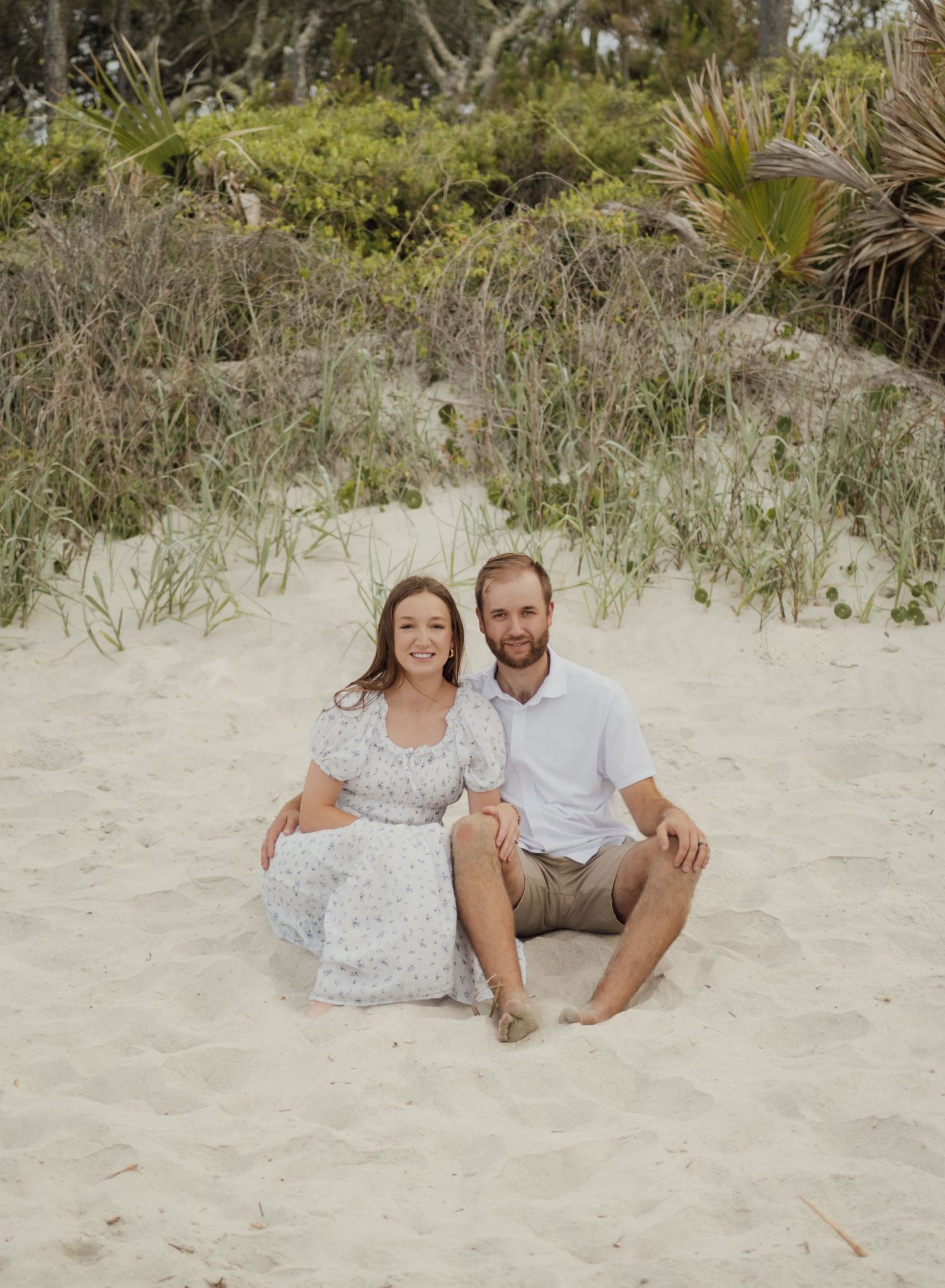 Couple sitting together on white sand beach dunes, woman in floral dress and man in white shirt, with lush greenery behind them.