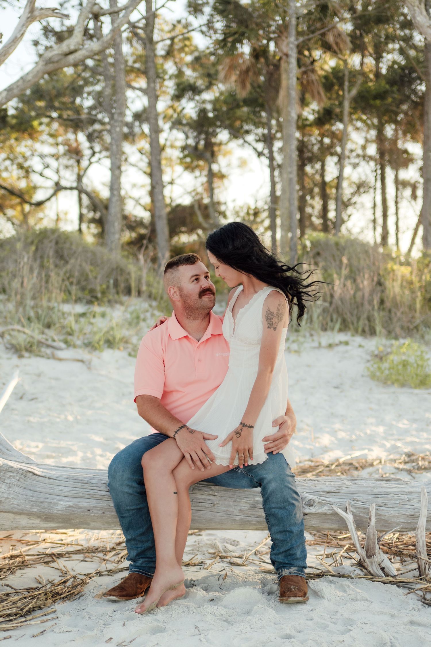 Couple sitting on driftwood at sandy beach, man in pink shirt and jeans, woman in white dress, trees in background.