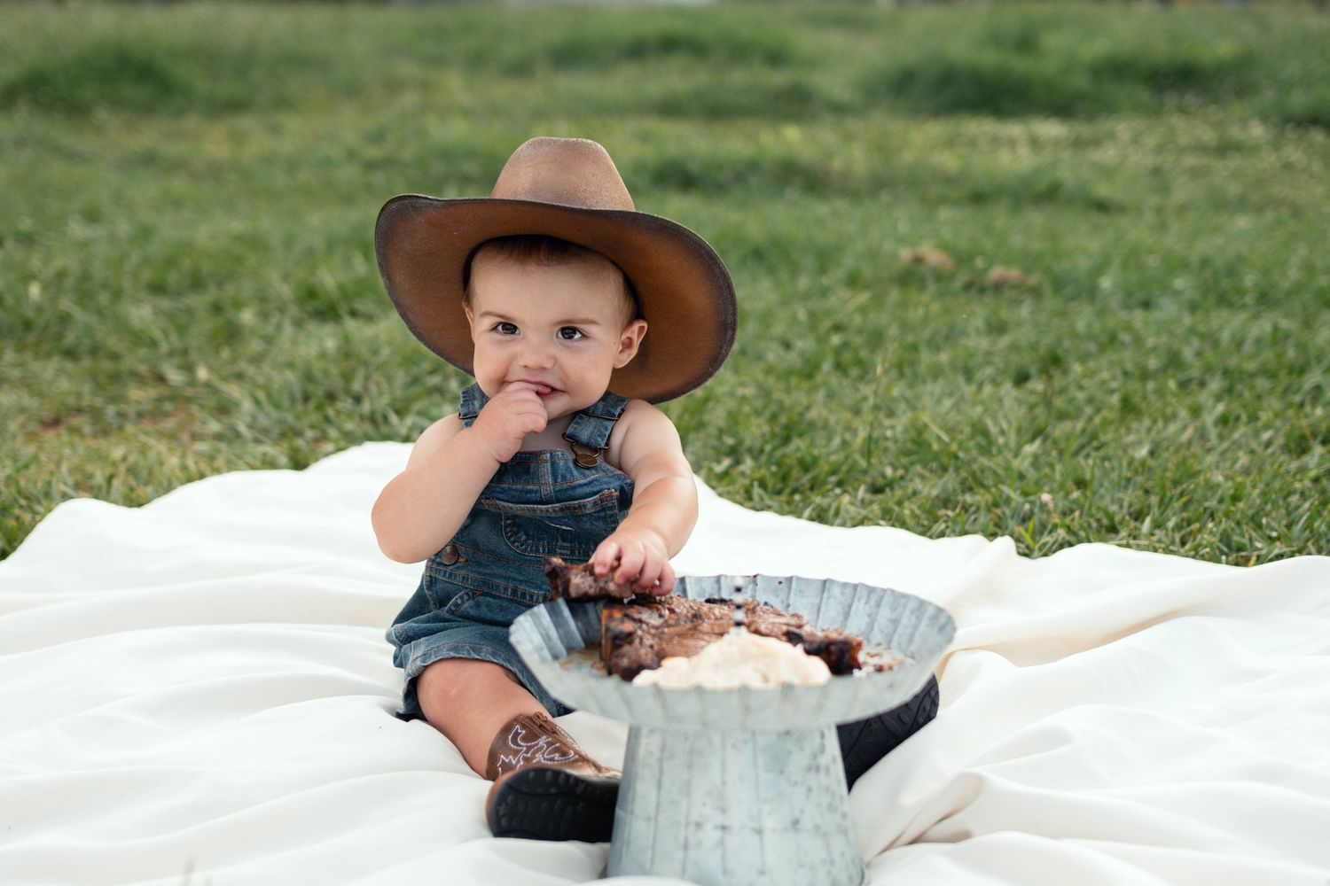 Baby in cowboy hat sitting on white blanket outdoors, enjoying cake smash with galvanized tub on green grass.