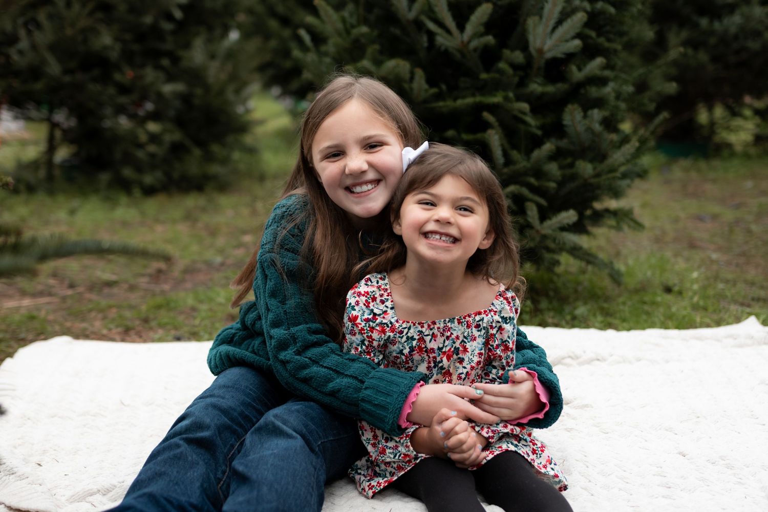 Two smiling sisters hugging on a white blanket at a Christmas tree farm, older girl in green sweater.