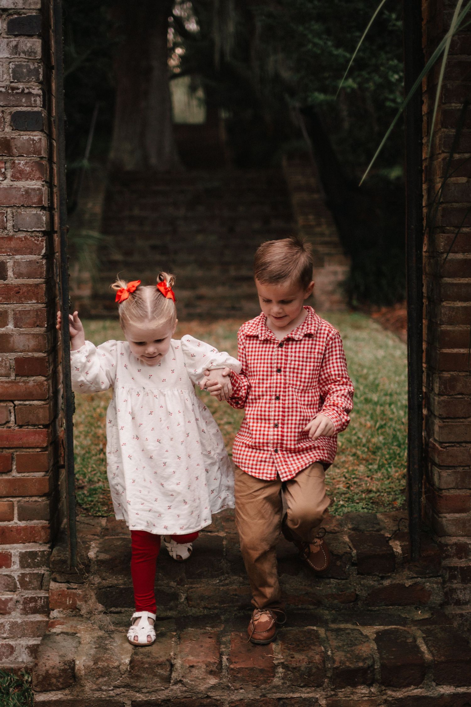 Two young children, a girl in white dress with red tights and a boy in plaid shirt, walk hand-in-hand through a brick archway.