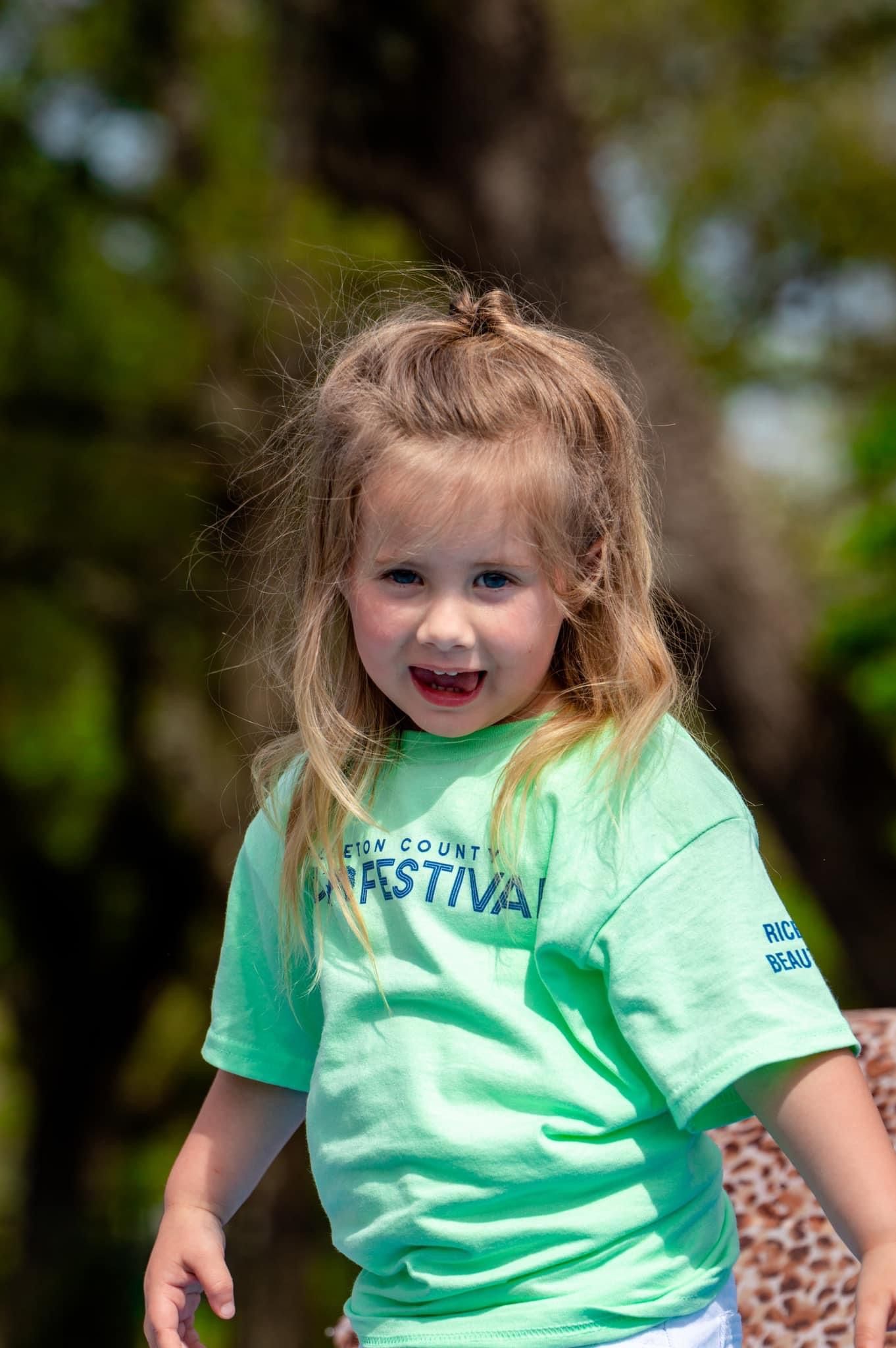 A smiling blonde girl in a mint green festival t-shirt poses outdoors with trees in the background.