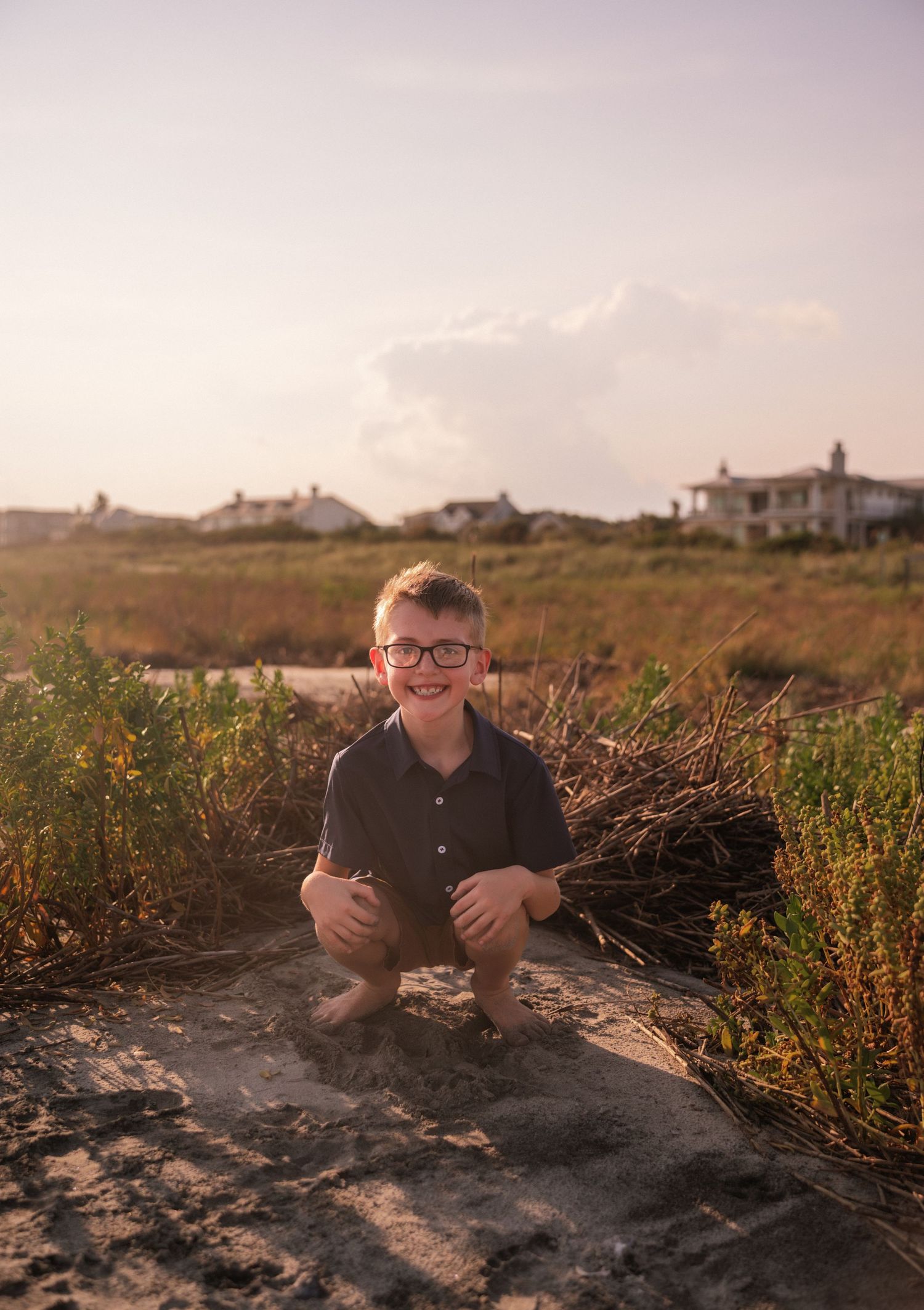 A smiling boy with glasses crouches on sandy beach dunes with coastal homes and golden grass in the background.