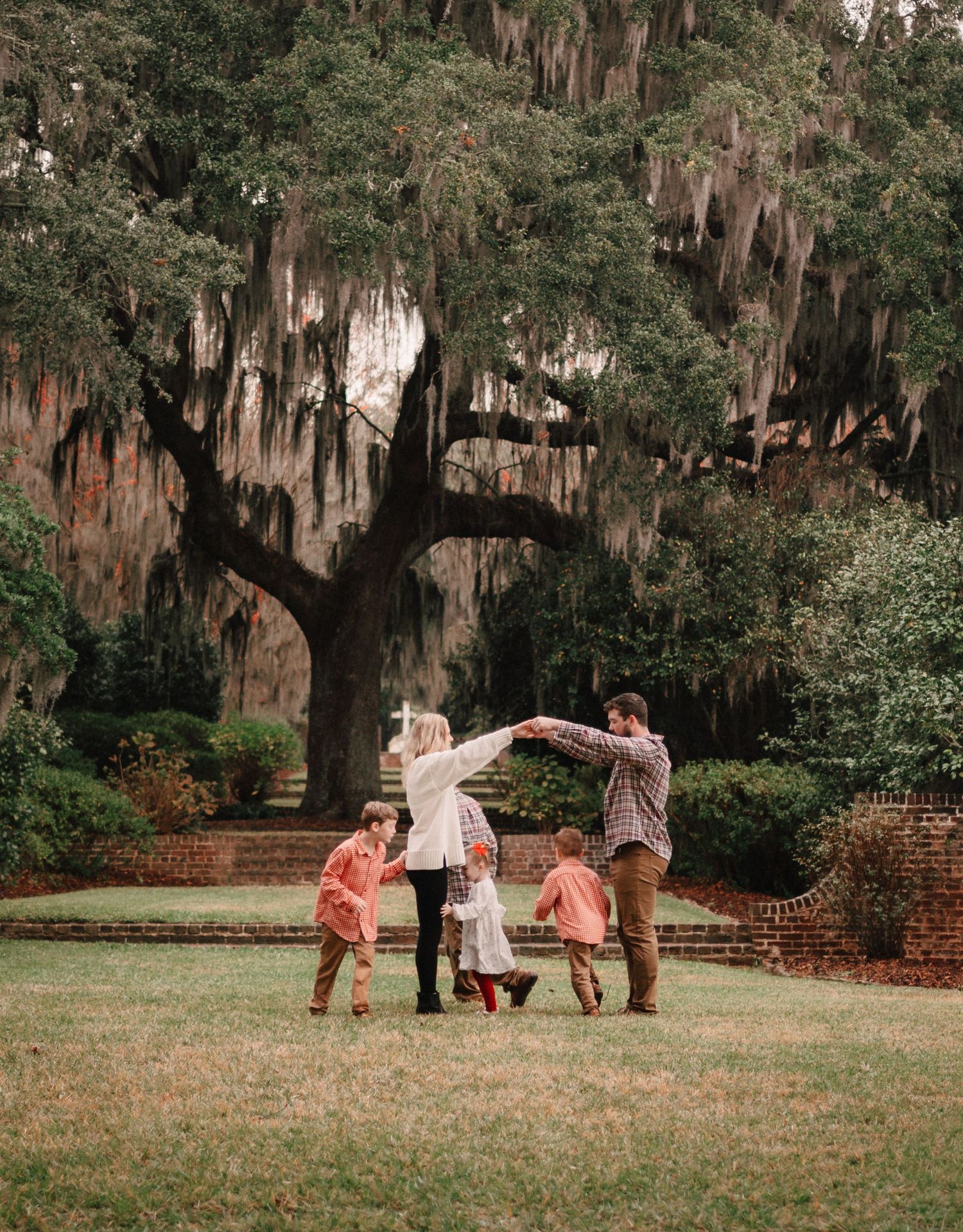 Family dancing together under a large moss-draped oak tree on a green lawn with children joining hands.