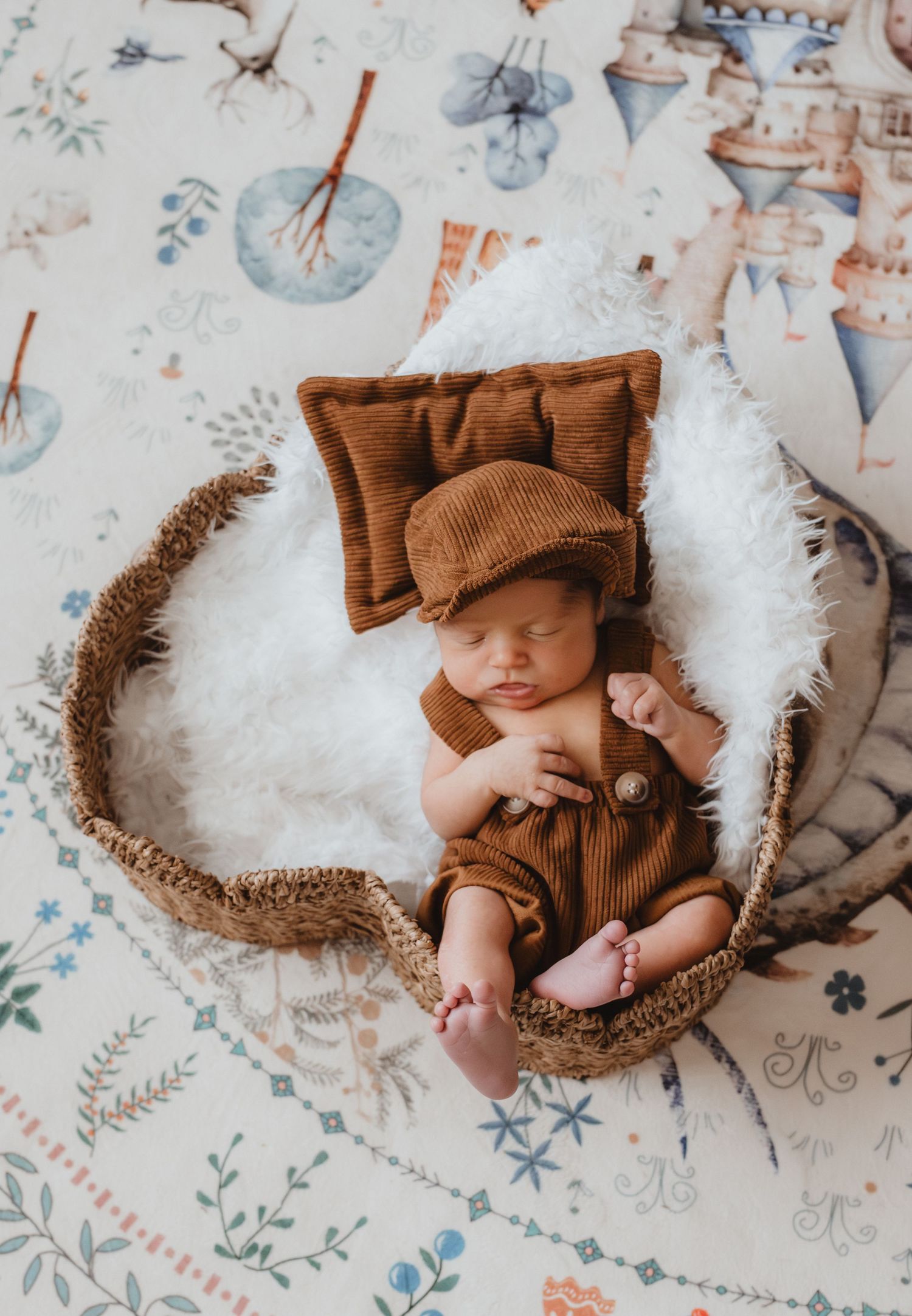 Sleeping newborn baby in brown outfit and newsboy cap resting in a wicker basket with white fur and floral backdrop.